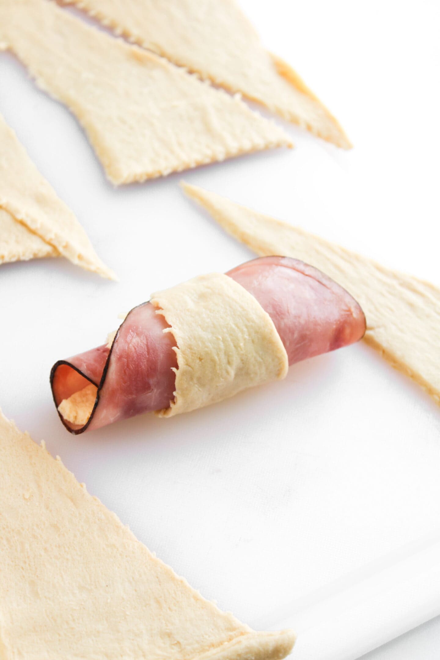 A slice of ham is wrapped in uncooked crescent roll dough on a white surface, preparing to make delicious Ham and Cheese Crescent Rolls, with more dough pieces nearby.