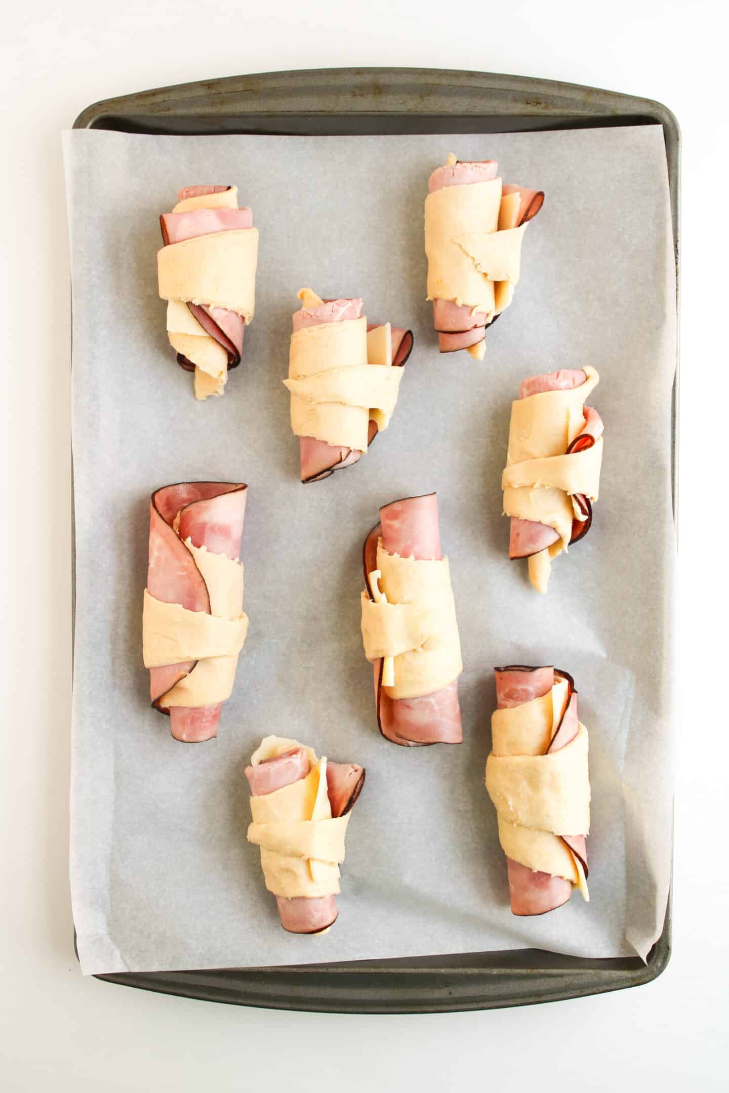 A baking tray lined with parchment paper holds seven unbaked Ham and Cheese Crescent Rolls, each roll wrapped around savory slices of ham.