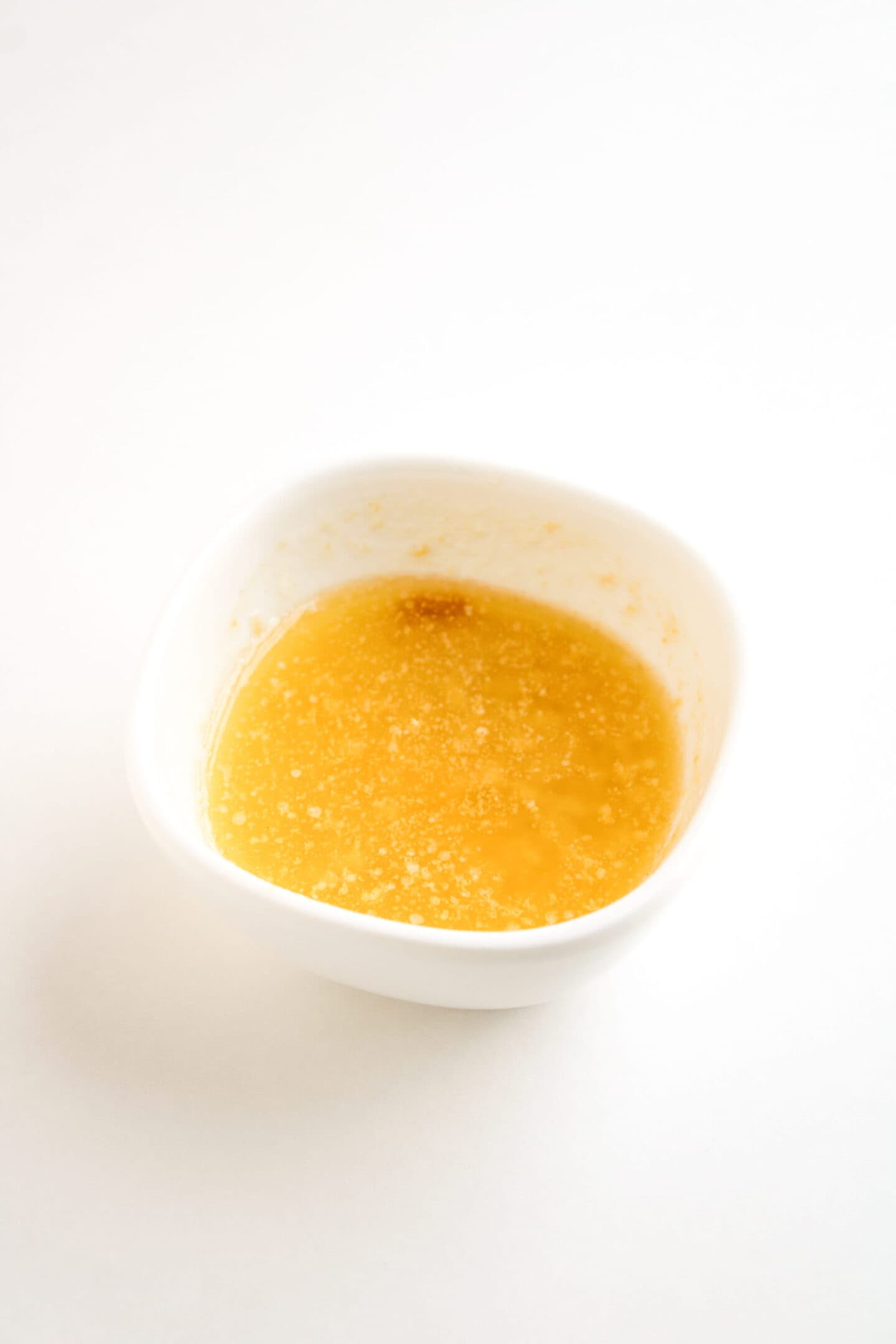 A small white bowl containing melted and slightly separated butter on a white surface, perfect for brushing over freshly baked Ham and Cheese Crescent Rolls.