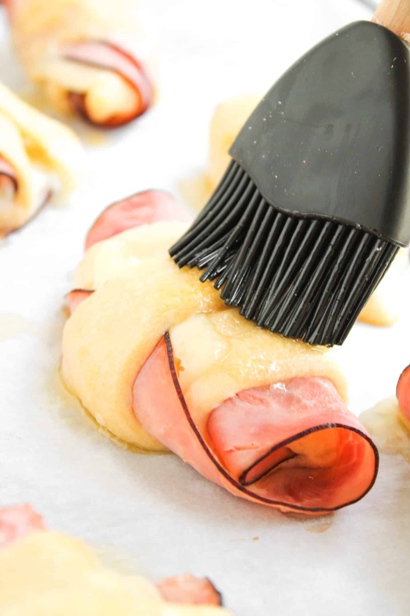 A pastry brush coats an unbaked Ham and Cheese Crescent Roll with a yellow glaze on a baking sheet.