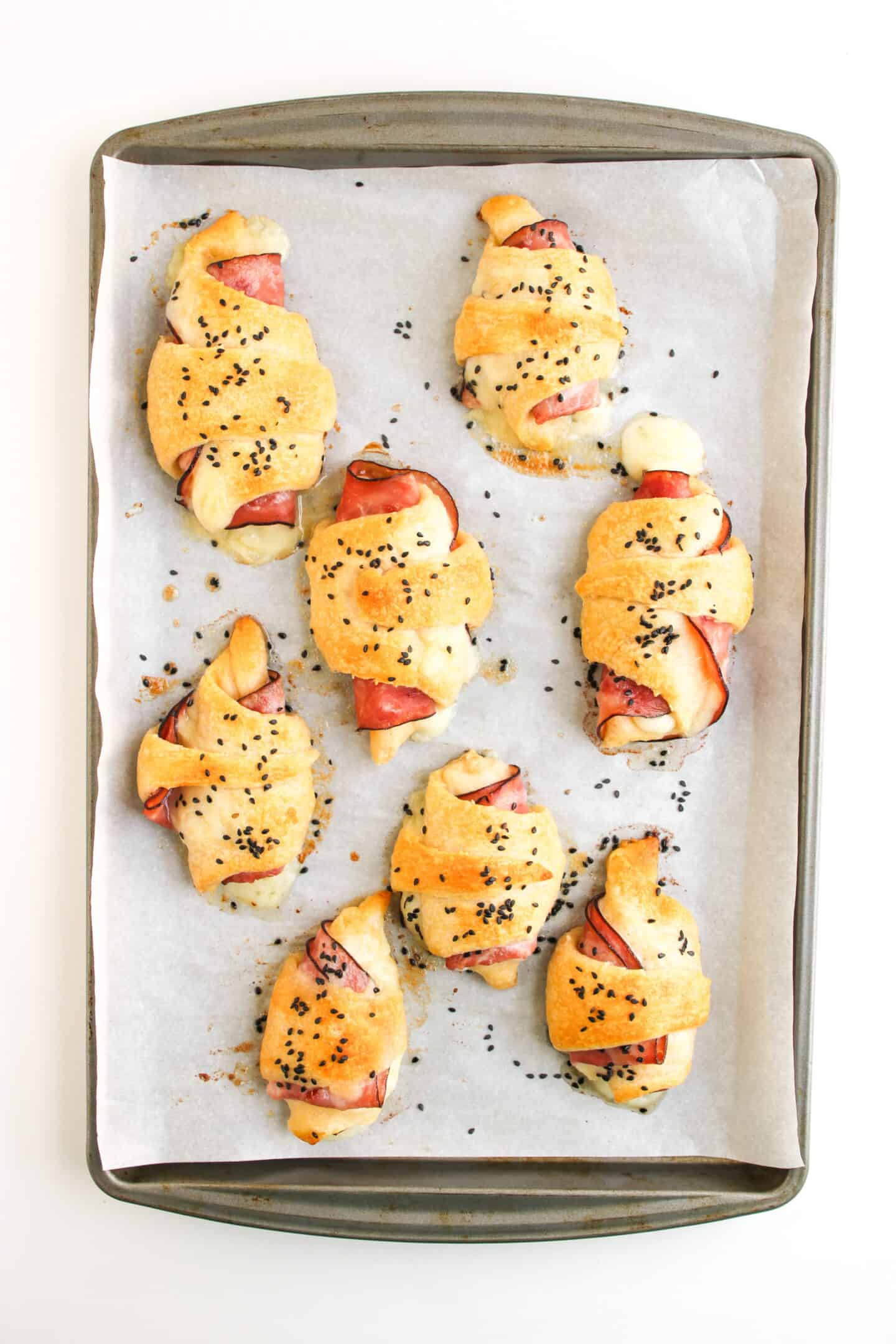 A baking tray lined with parchment paper holds seven Ham and Cheese Crescent Rolls, each golden, filled with savory ham and cheese, and topped with black sesame seeds.