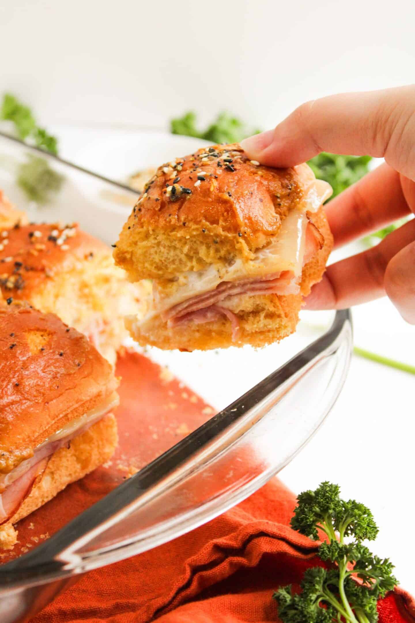 A hand holds a ham and cheese slider with a golden bun, lifted from a glass baking dish filled with more sliders—perfect Easter Party Food, with fresh parsley visible nearby.