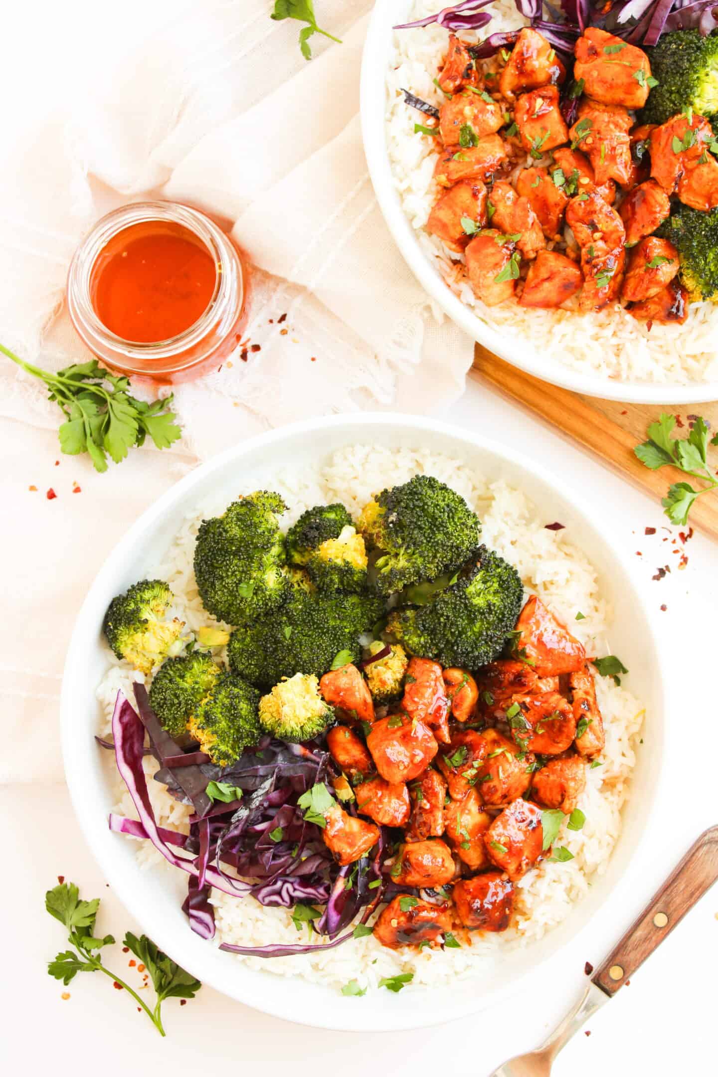 A bowl of white rice topped with broccoli, red cabbage, and tender Hot Honey Chicken pieces, served with a jar of sauce and fresh herbs nearby.