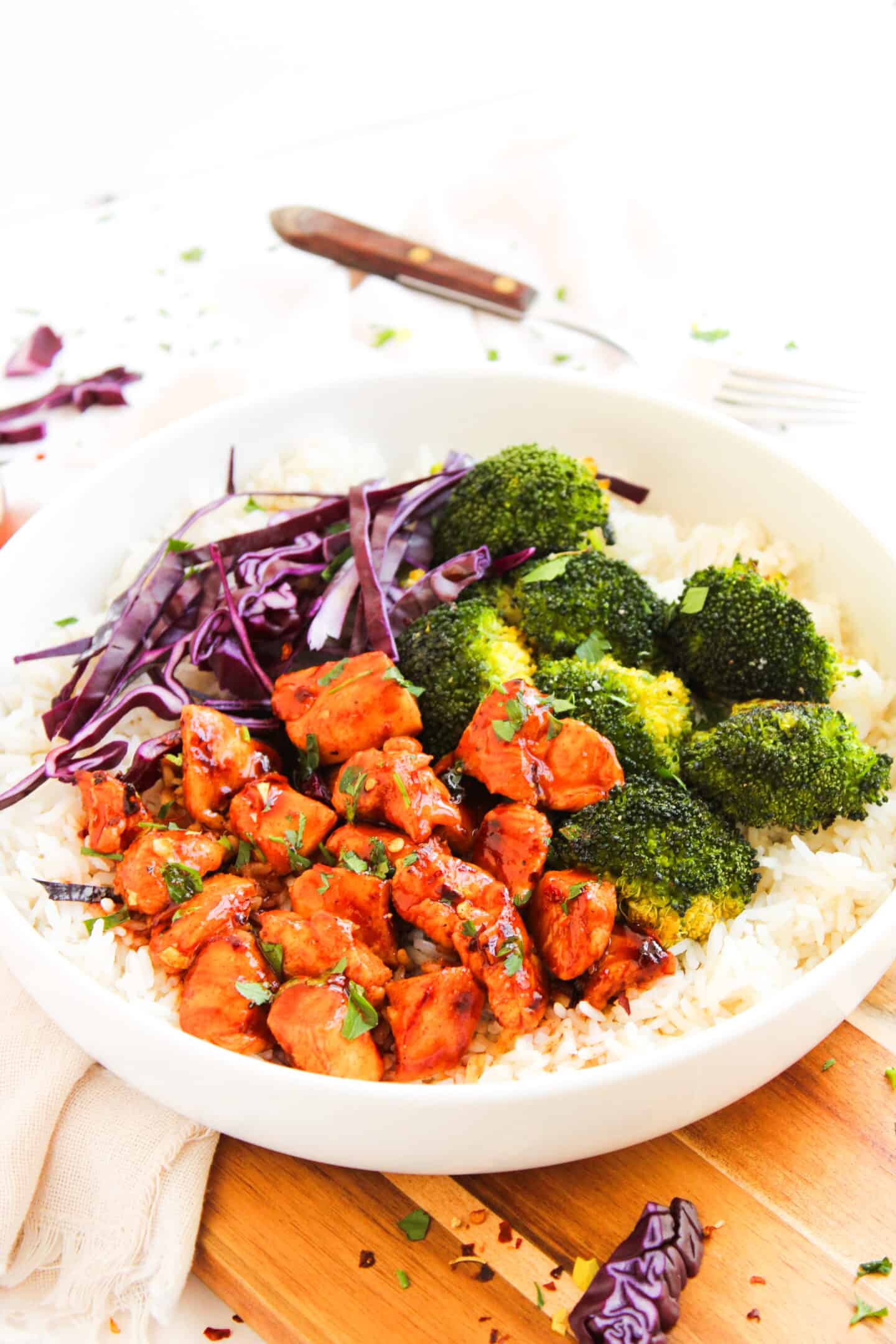A white bowl filled with white rice, cooked broccoli, shredded purple cabbage, and glazed Hot Honey Chicken pieces garnished with herbs—a perfect Hot Honey Chicken Bowl.