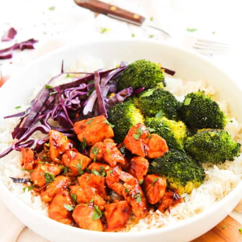 A white bowl filled with white rice, cooked broccoli, shredded purple cabbage, and glazed Hot Honey Chicken pieces garnished with herbs—a perfect Hot Honey Chicken Bowl.