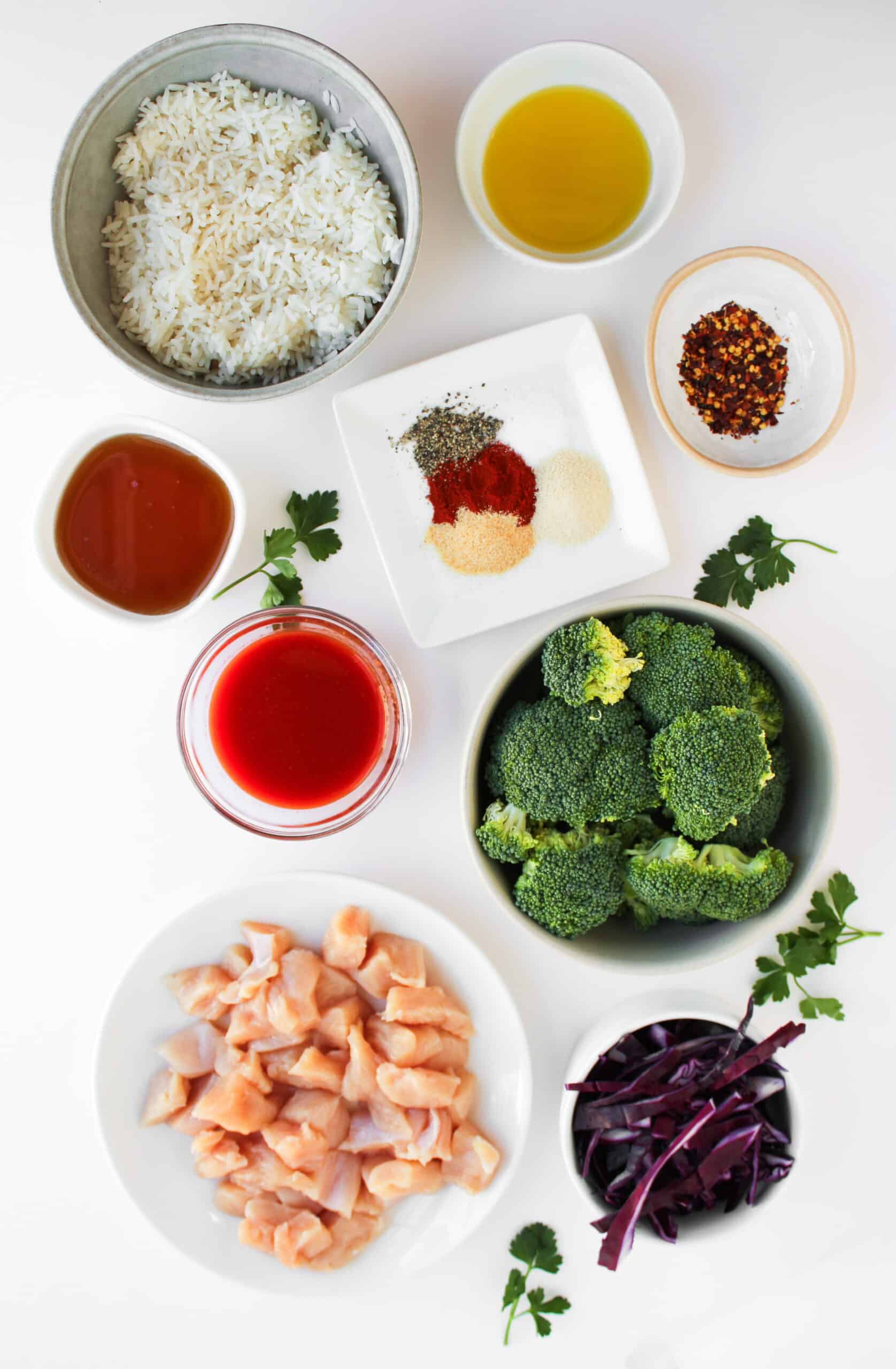 Overhead view of bowls containing uncooked rice, olive oil, red pepper flakes, seasonings, soy sauce, tomato sauce, broccoli florets, chopped raw chicken for a Chicken Bowl, shredded red cabbage—perfect for a Hot Honey Chicken recipe.