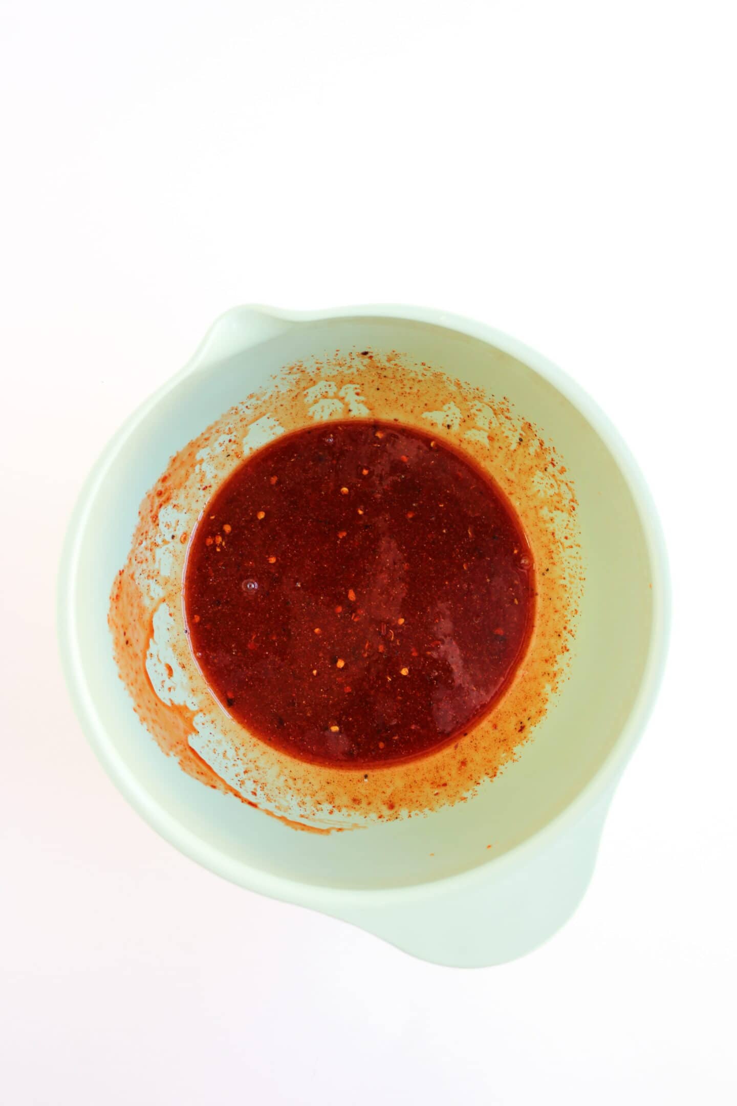 A white mixing bowl containing a thick, red sauce with visible spices and seeds, perfect for drizzling over a Hot Honey Chicken Bowl, set against a white background.