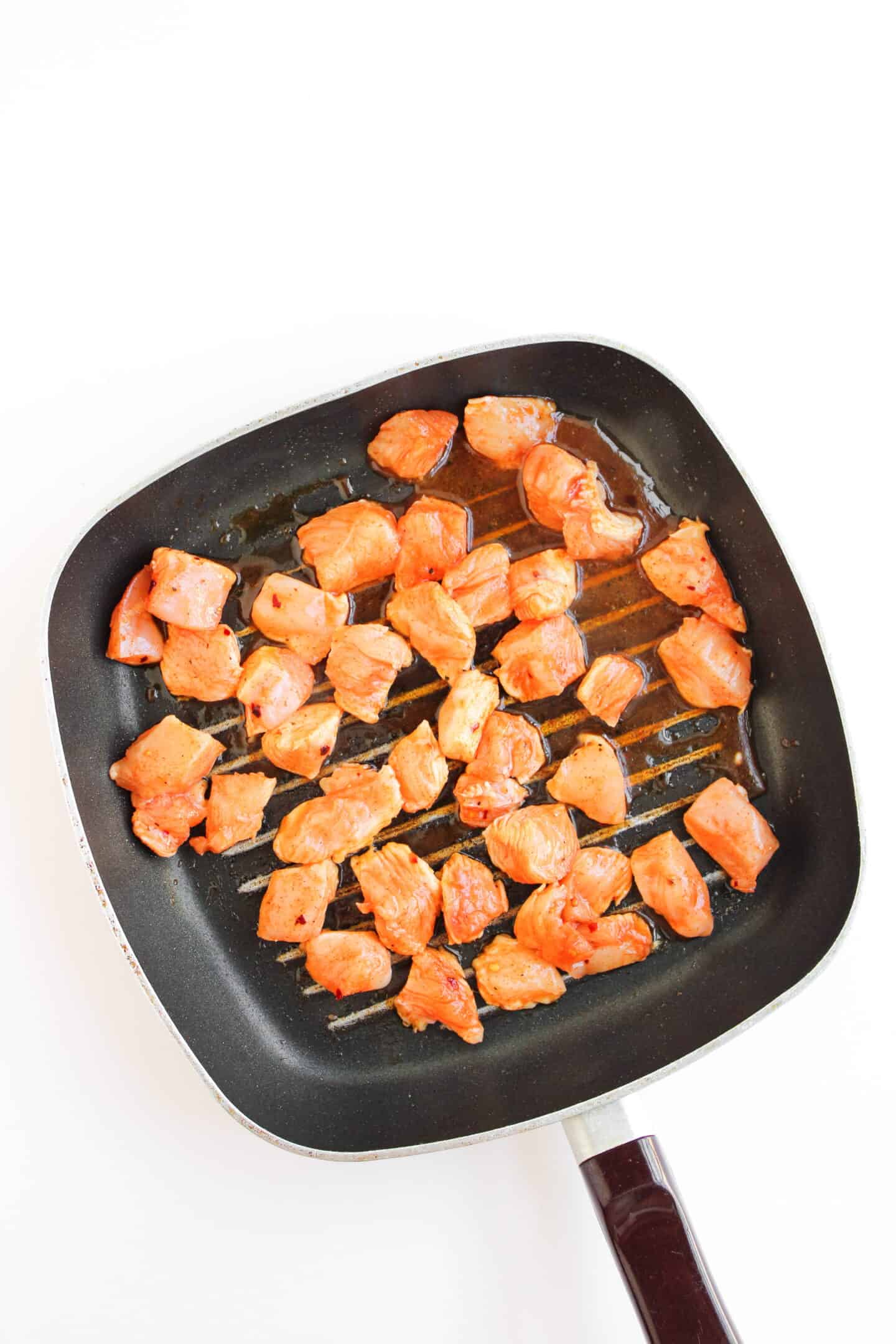 Cubed pieces of seasoned hot honey chicken are cooking in a black grill pan against a white background.