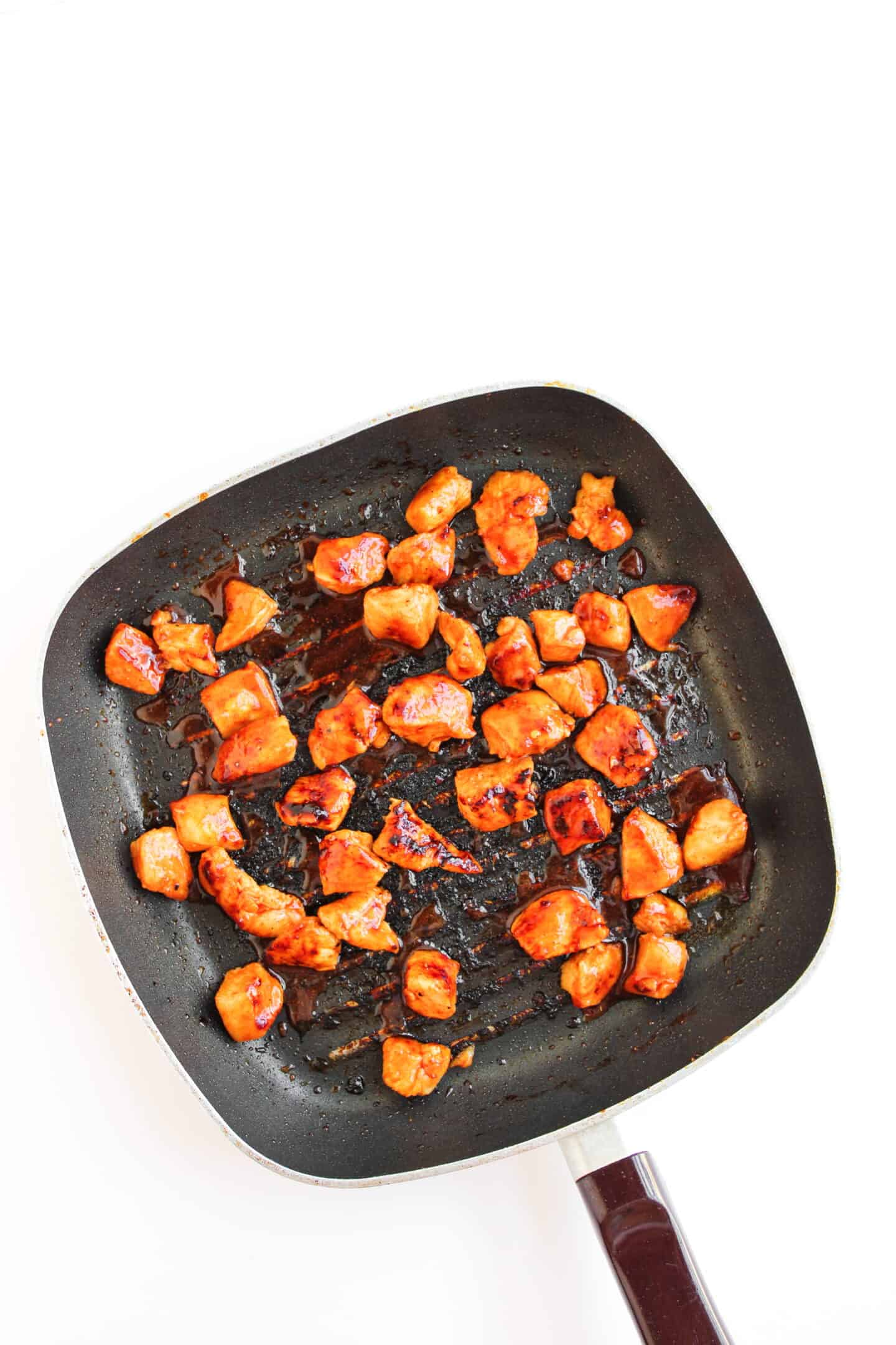 Cubed pieces of cooked, browned chicken coated in hot honey sauce make this Hot Honey Chicken Bowl a flavorful delight, served in a black square frying pan on a white background.