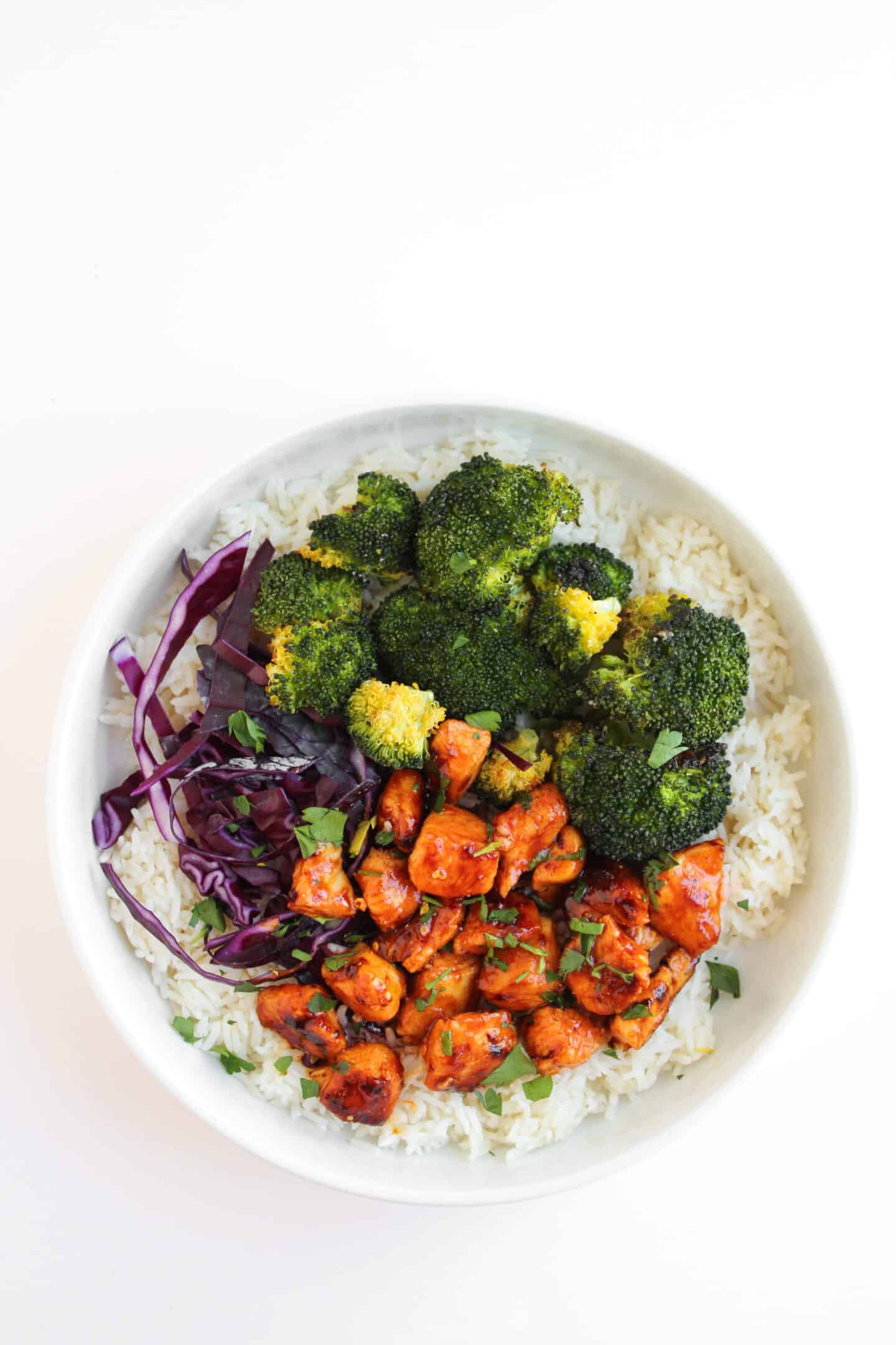 A white bowl containing white rice, roasted broccoli, red cabbage, and diced Hot Honey Chicken, garnished with chopped herbs—a flavorful Hot Honey Chicken Bowl.