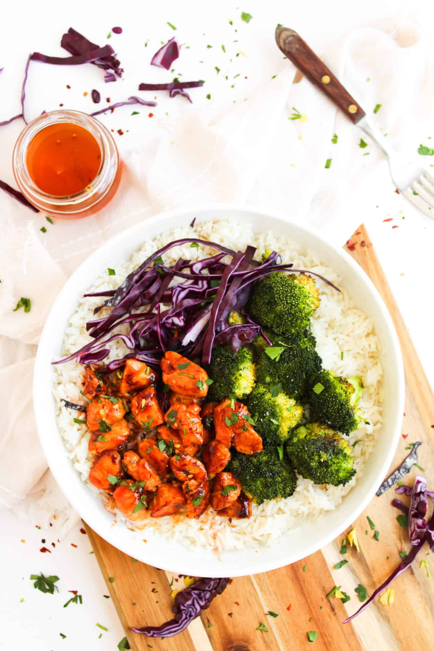 A Chicken Bowl filled with rice, broccoli, shredded red cabbage, and saucy chicken pieces sits on a wooden board beside a jar of sauce and a fork.