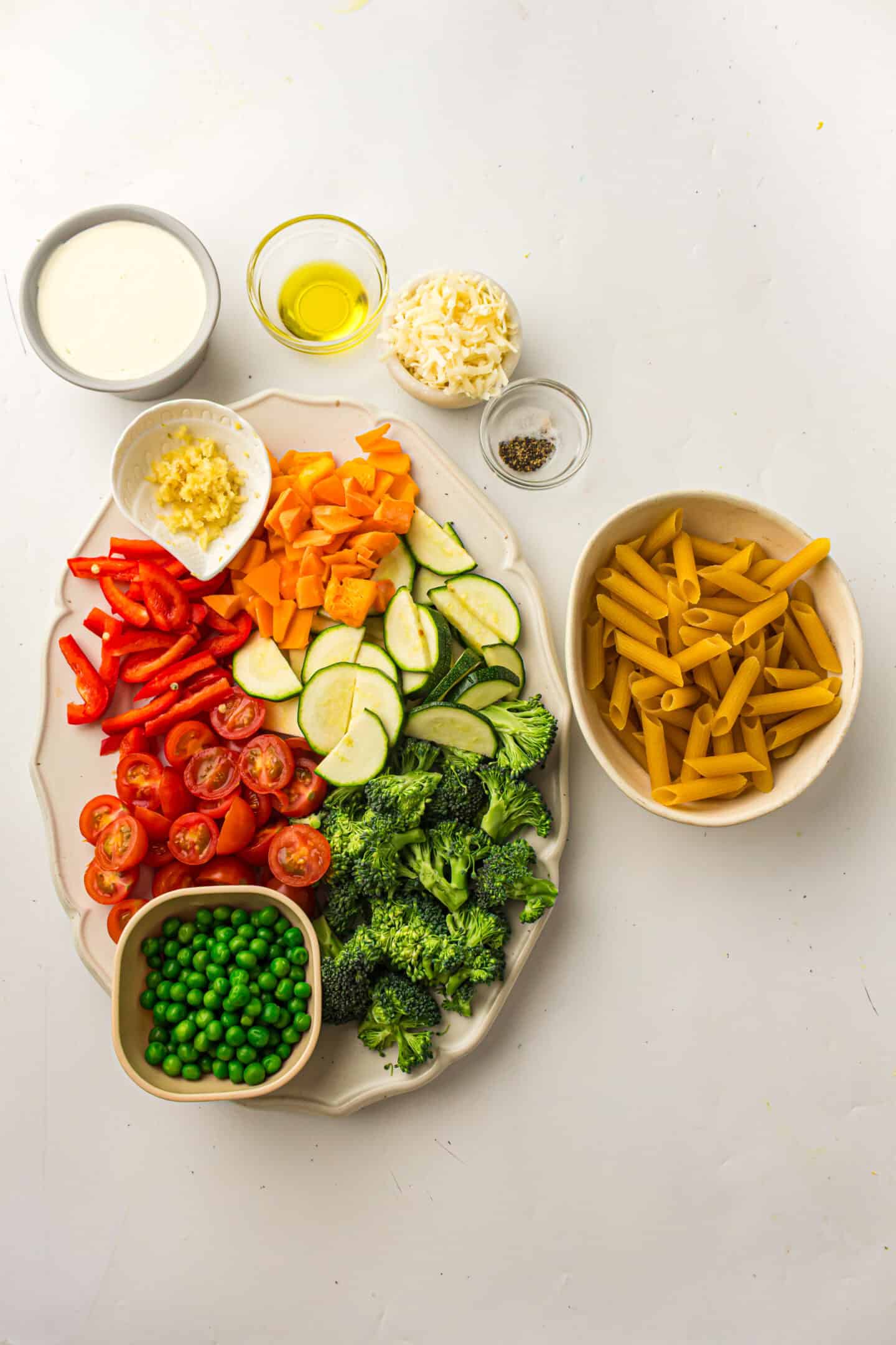 A platter with chopped red bell pepper, sweet potato, zucchini, cherry tomatoes, broccoli, and peas sits next to bowls of dry penne and toppings—perfect ingredients for a fresh vegetable pasta or classic Pasta Primavera.