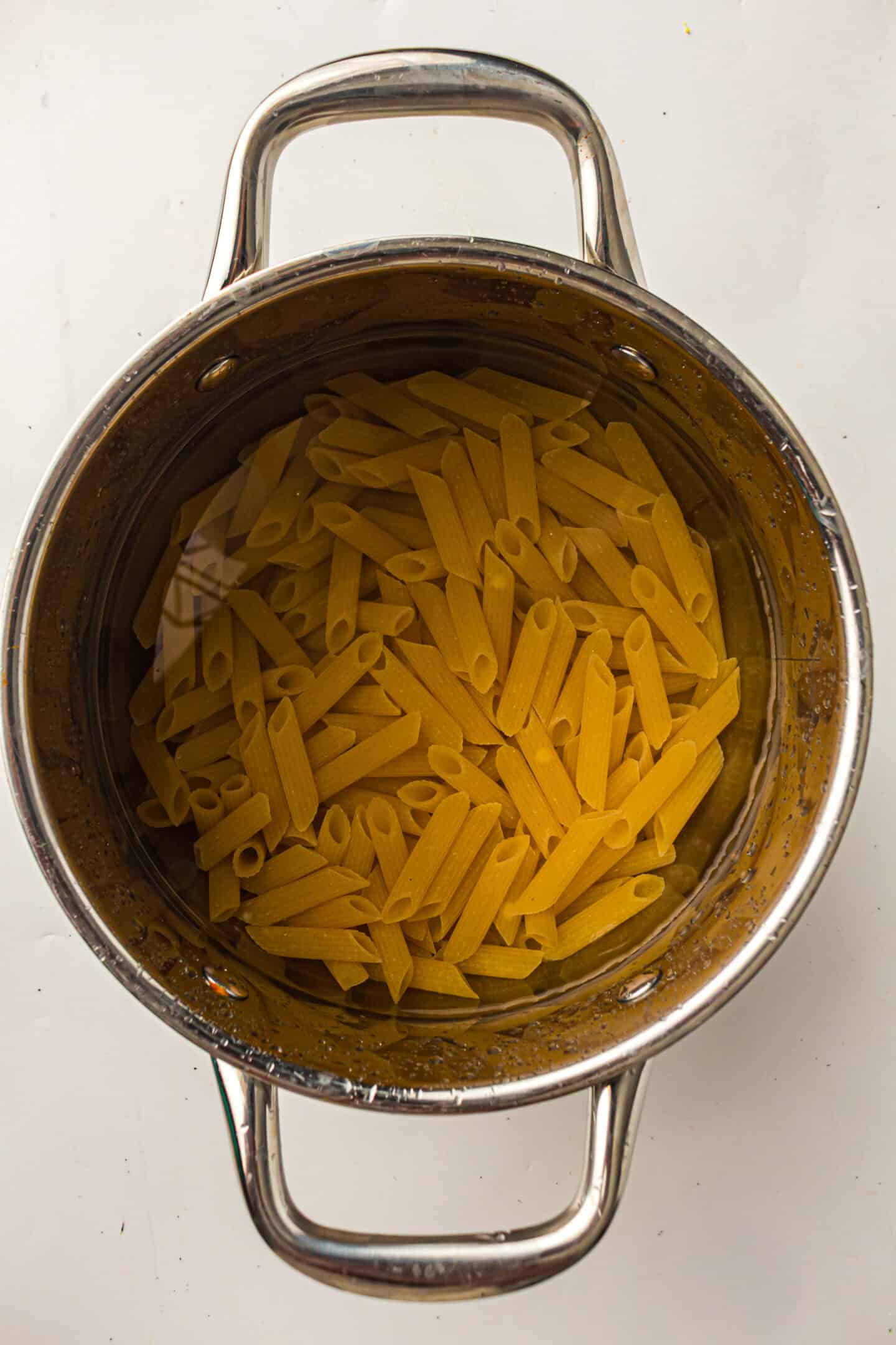 A stainless steel pot filled with uncooked penne pasta and water, viewed from above on a white surface—perfect for starting your favorite Pasta Primavera or vegetarian pasta recipe.