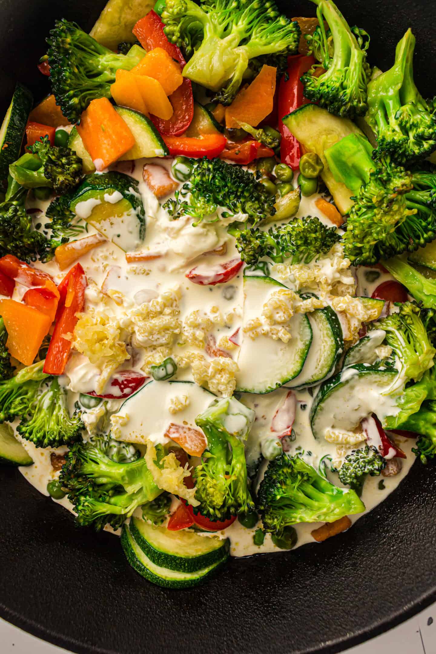 A close-up of assorted vegetables, including broccoli, zucchini, carrots, and peppers, in a pan with cream sauce being poured over them—a delicious base for a vegetarian pasta or classic Pasta Primavera recipe.