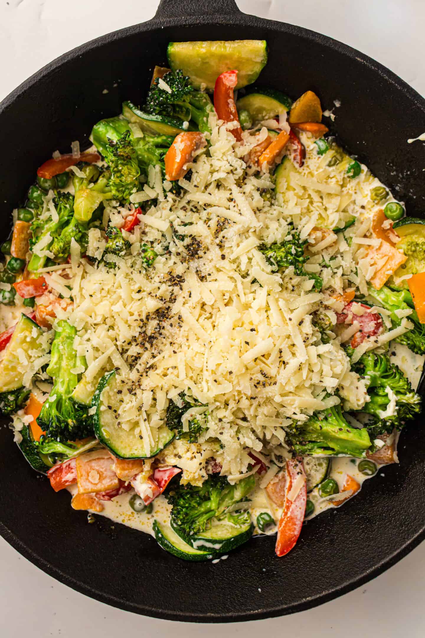 A skillet filled with sautéed vegetables, including broccoli, zucchini, and bell peppers, creates a vibrant Pasta Primavera, topped with shredded cheese and cracked black pepper.