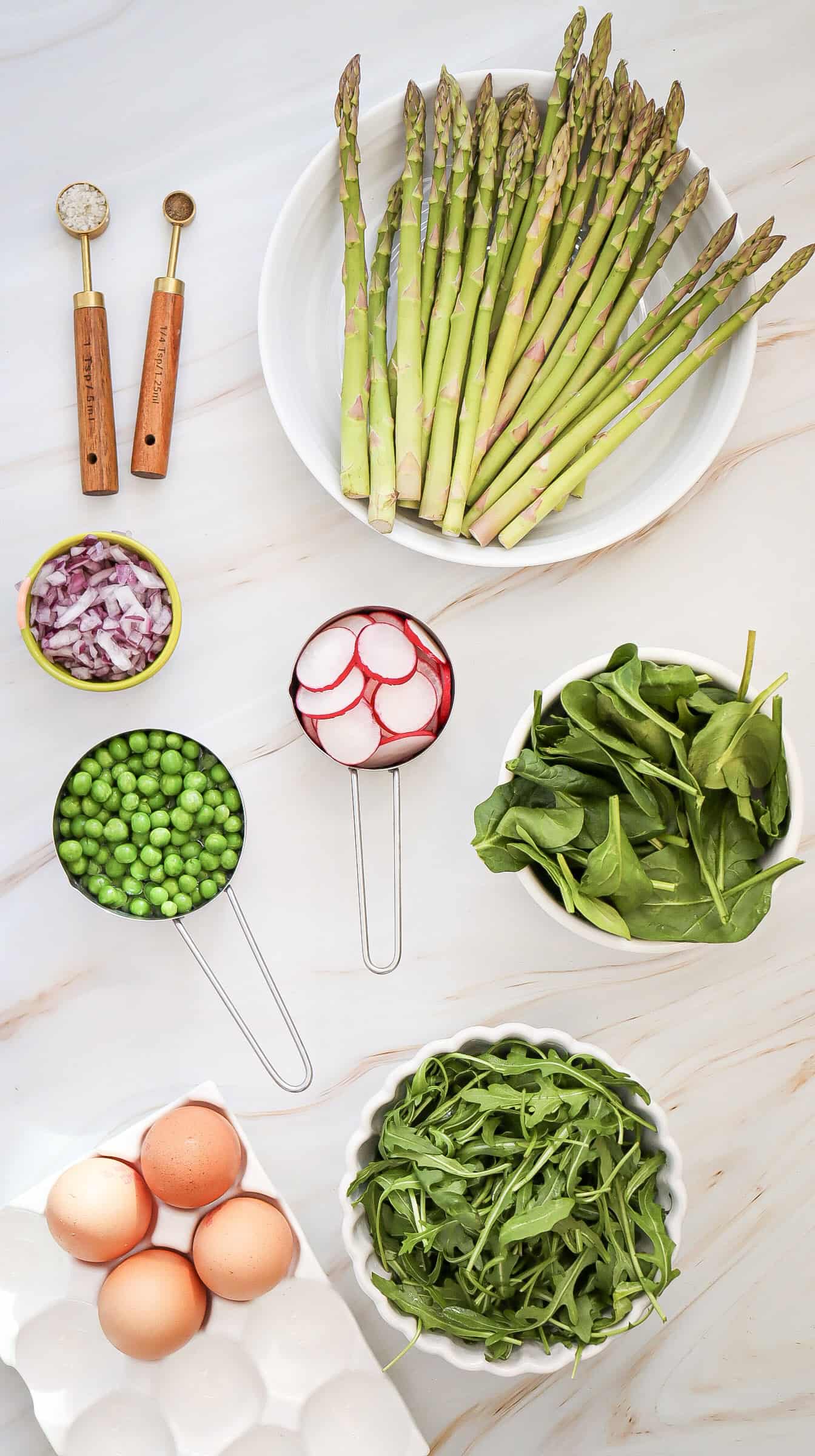 A flat lay of fresh ingredients including asparagus, red onion, radish, peas, spinach, arugula, and eggs arranged on a white surface.