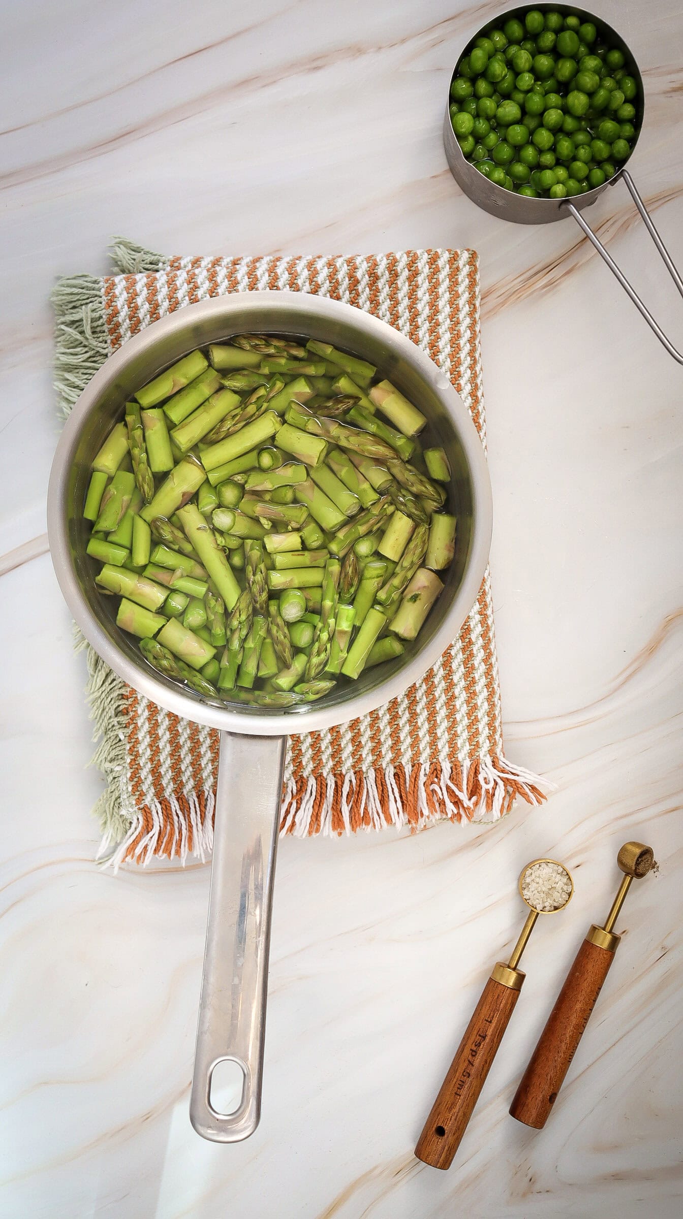 A saucepan with chopped asparagus in water sits on a cloth, next to a measuring cup with green peas—perfect ingredients for a fresh asparagus salad or vibrant pea salad—alongside two wooden-handled utensils on a marble countertop.