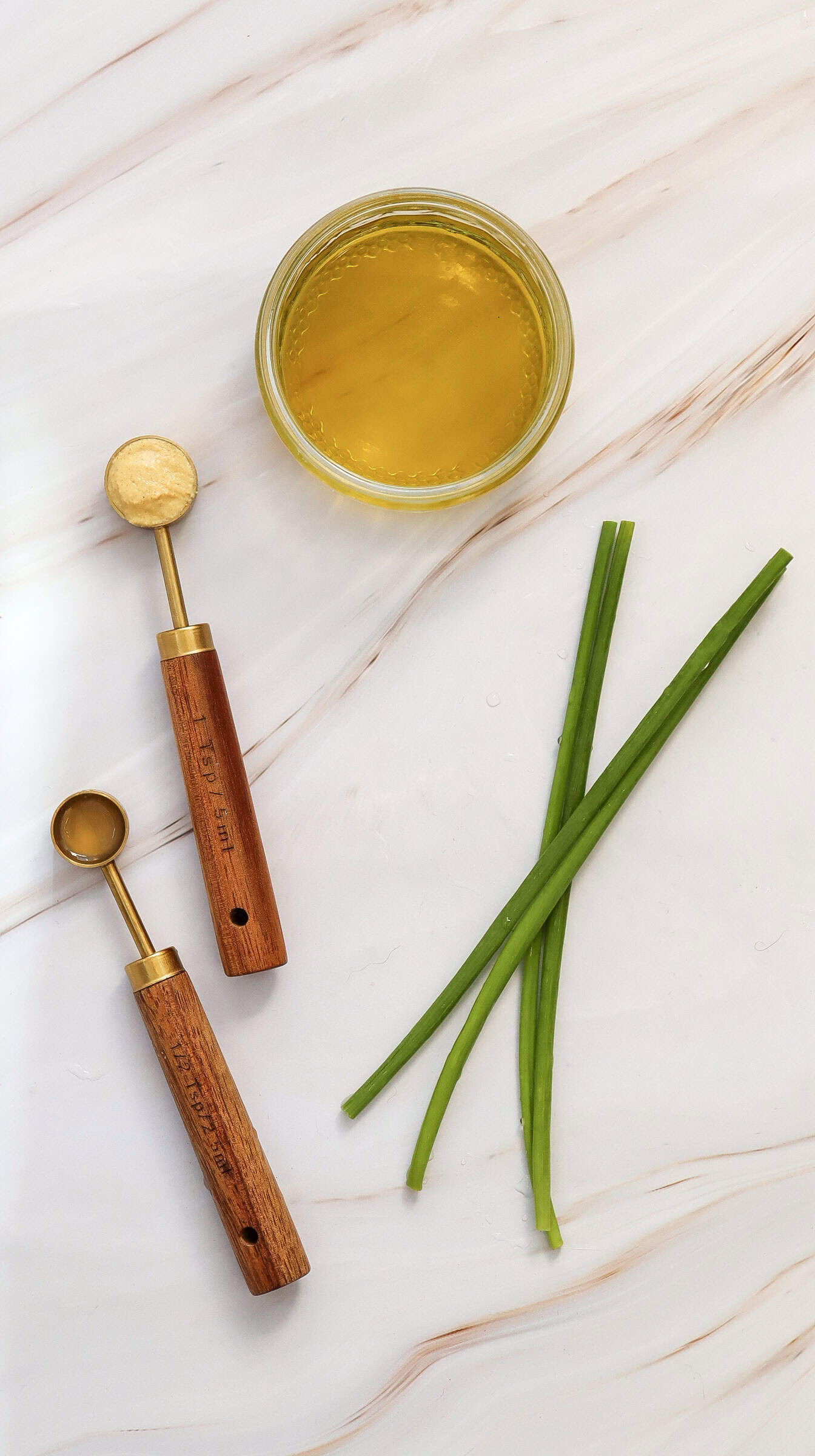 A small glass bowl of yellow lemon dressing, two melon ballers with wooden handles, and three chives sit on a white marble surface.