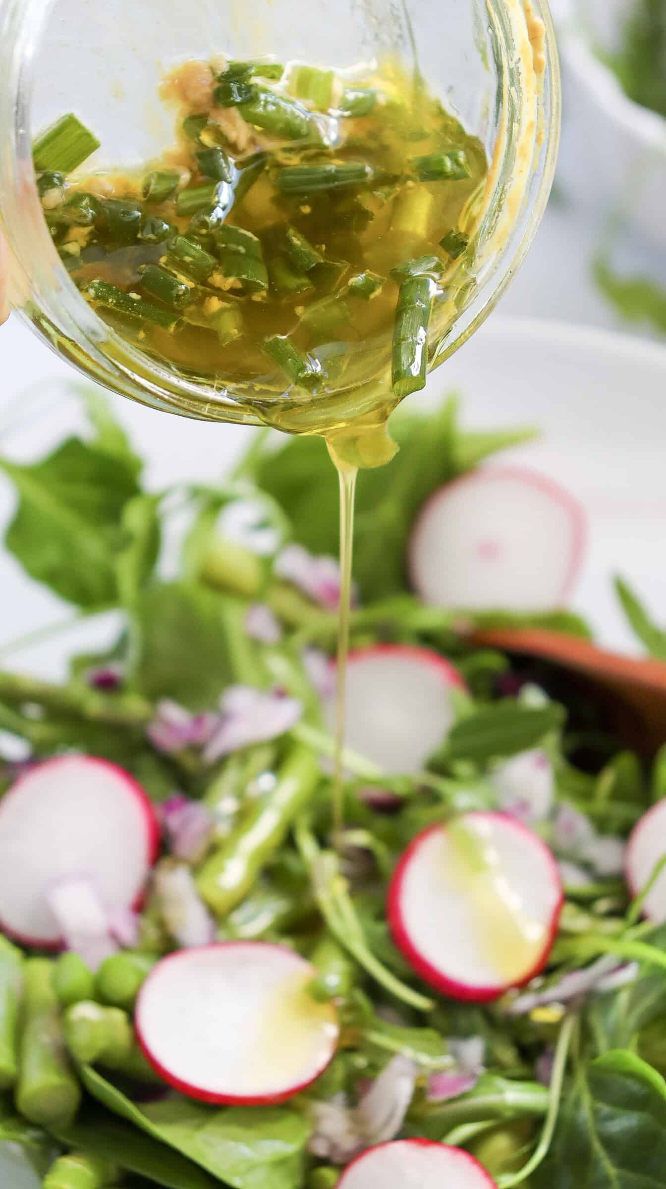 A close-up of a green pea salad with radish slices and asparagus, dressed with a vibrant yellow lemon dressing poured from a jar.