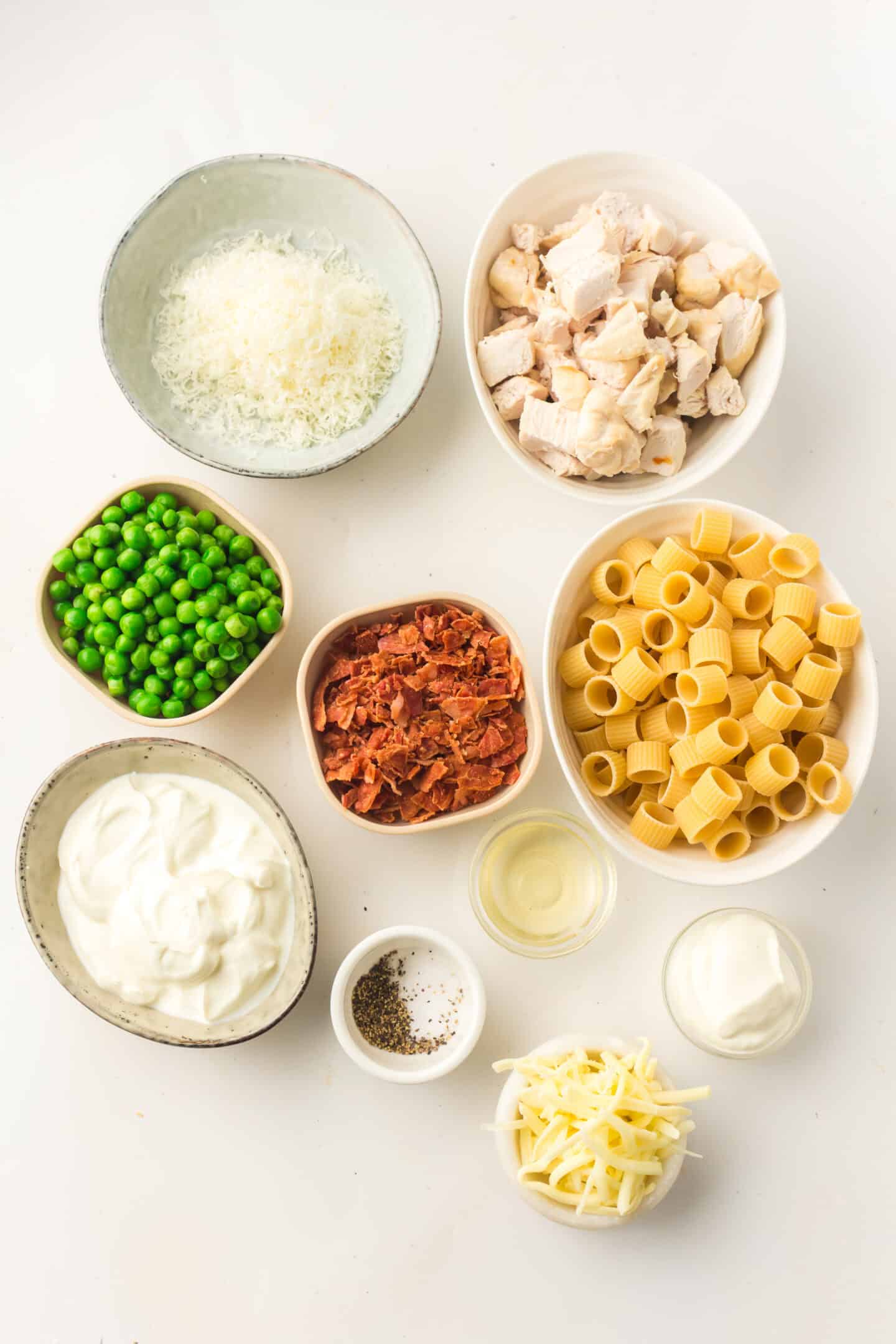Nine bowls containing ingredients for a Light Pasta Salad—grated cheese, cooked chicken pieces, green peas, cooked bacon, uncooked pasta, sour cream, cream, black pepper, and grated parmesan—are arranged on a white surface.