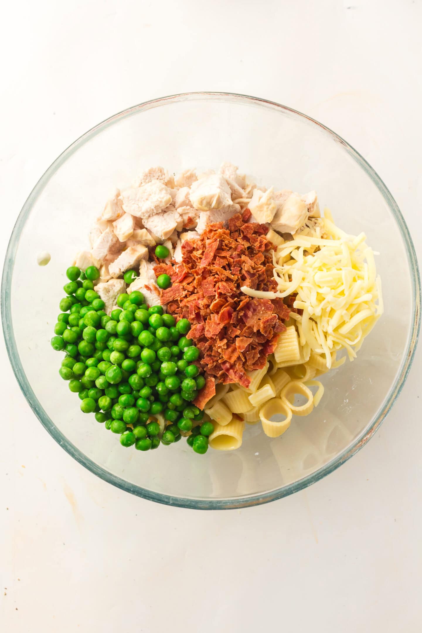 A glass bowl containing Chicken Pasta Salad with green peas, shredded cheese, diced cooked chicken, and crumbled bacon, all arranged in separate sections.