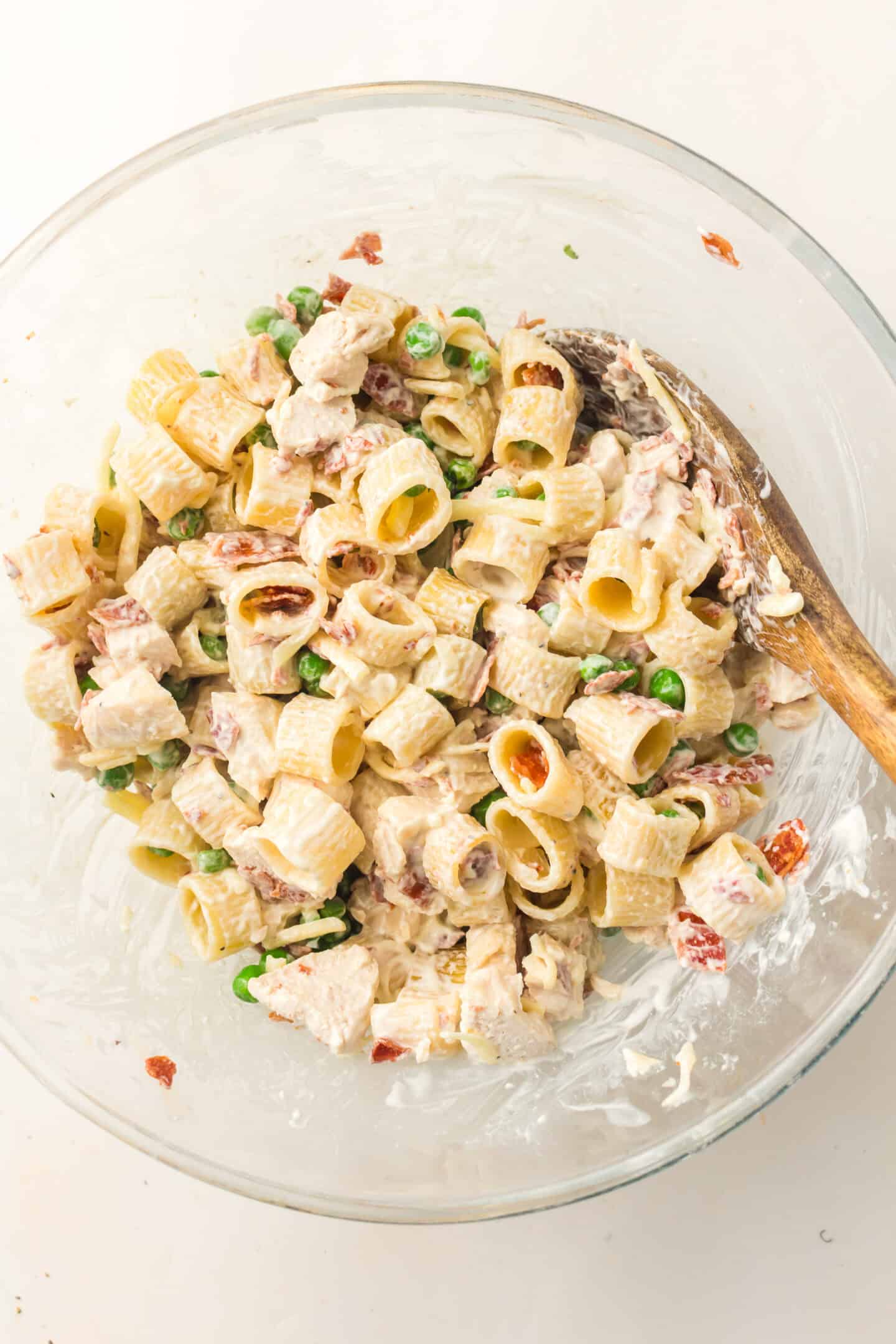 A glass bowl filled with creamy Chicken Pasta Salad featuring tube-shaped pasta, tender chicken pieces, peas, and sun-dried tomatoes, all mixed together with a wooden spoon.