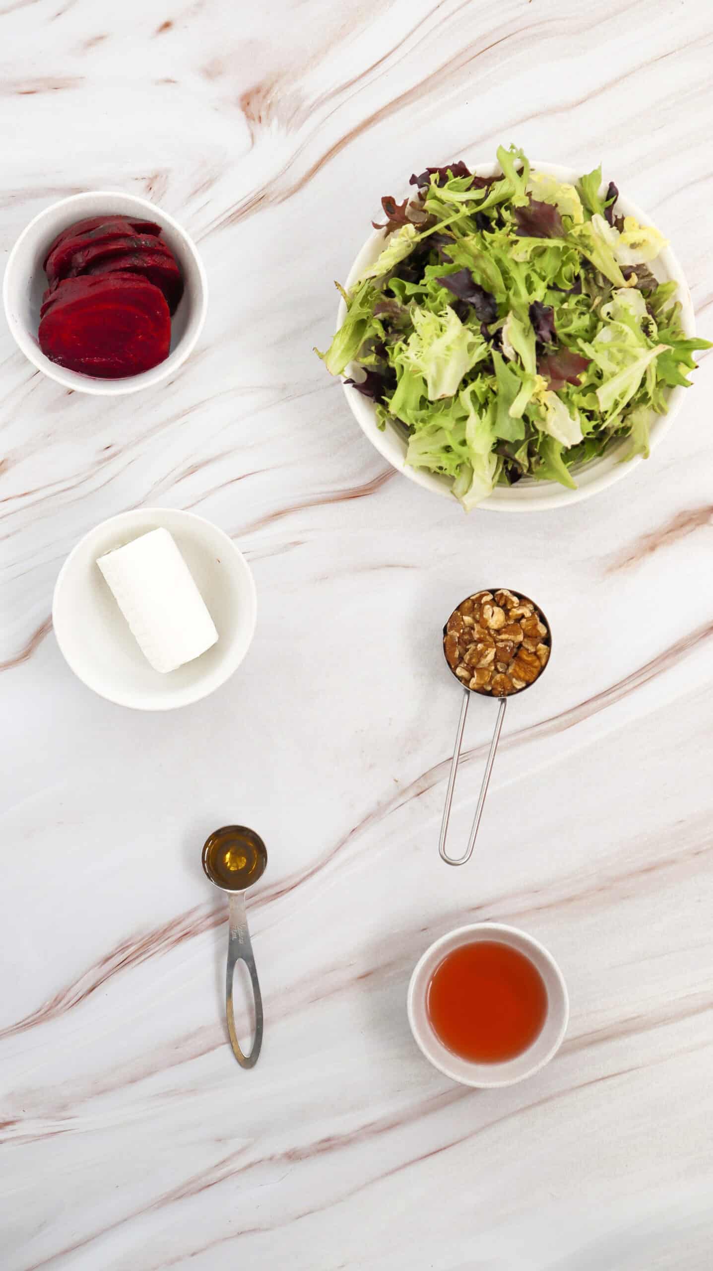 Overhead view of a healthy salad spread on marble: mixed greens, sliced beets, goat cheese, chopped nuts, honey, and vinaigrette in separate containers—perfect for assembling your own delicious goat cheese salad.
