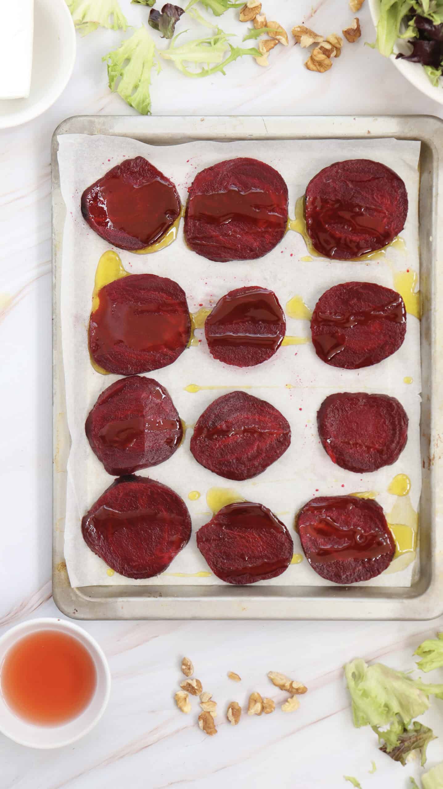 Sliced roasted beets on a parchment-lined baking sheet drizzled with olive oil, surrounded by salad greens, walnuts, and a bowl of vinaigrette—perfect for a vibrant grilled beetroot salad or a delightful walnut salad.
