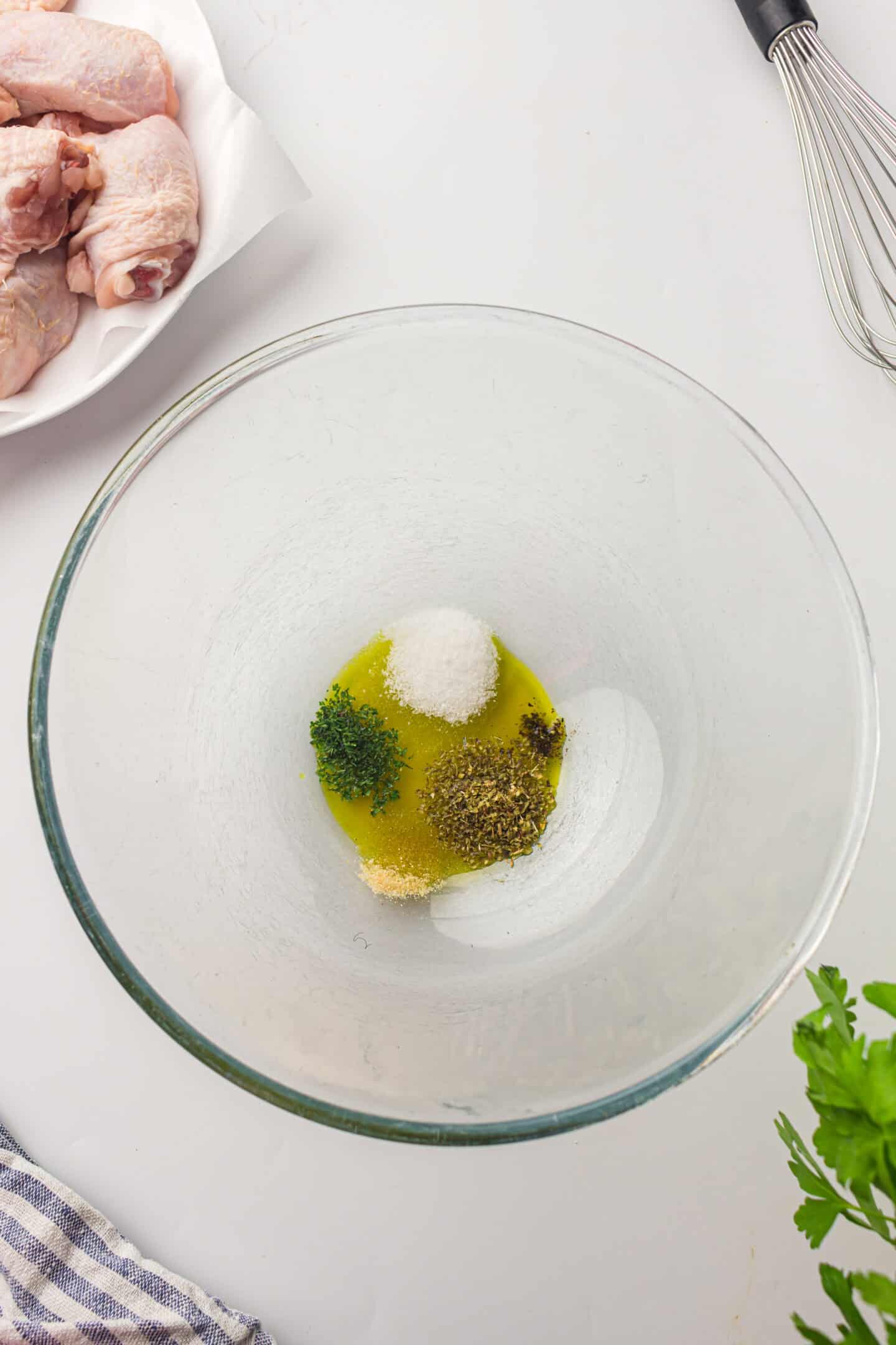A glass bowl with olive oil, salt, dried herbs, and chopped parsley sits on a white surface next to raw chicken thighs, a whisk, fresh parsley—perfect for prepping air fryer chicken.