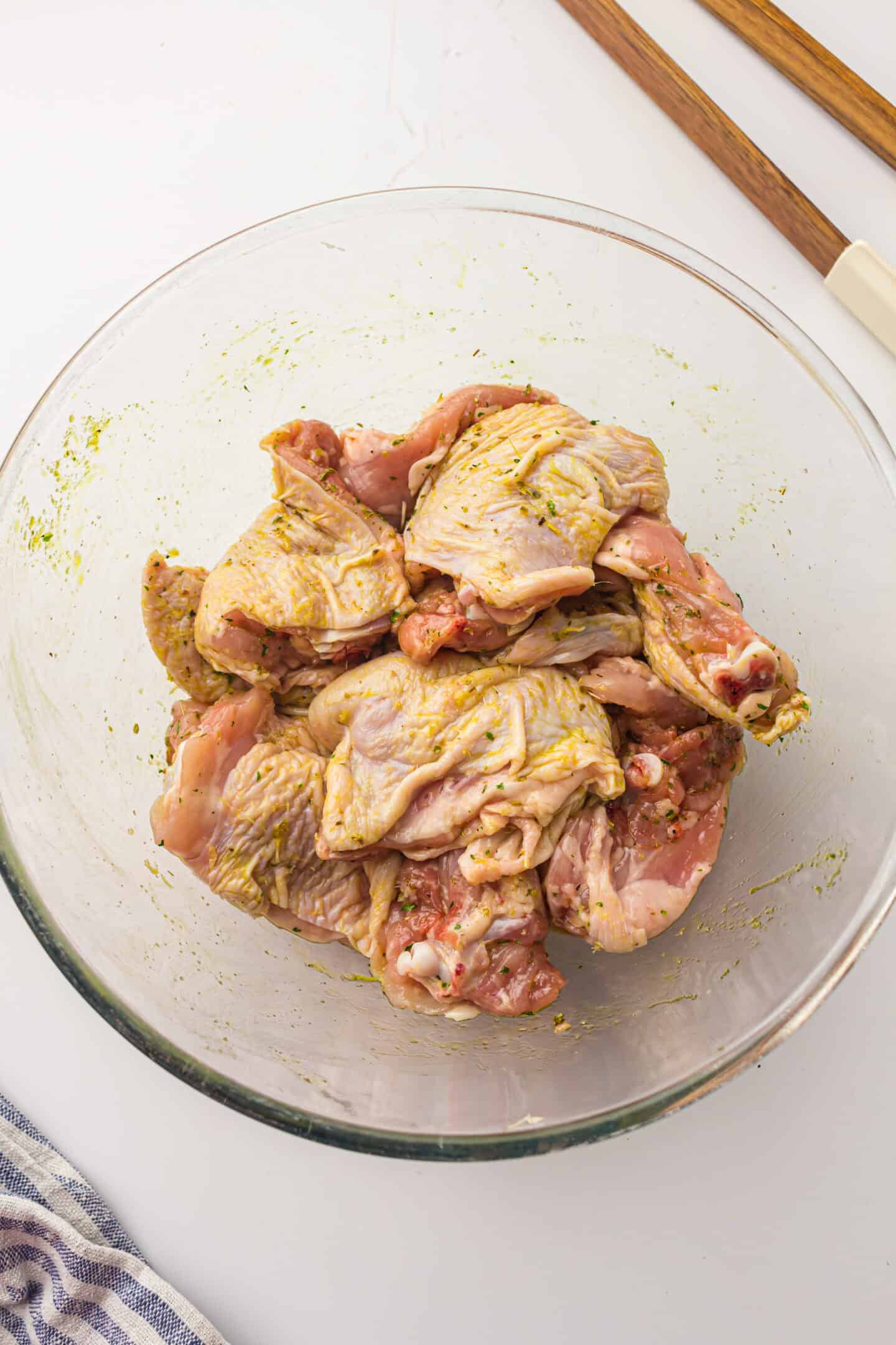 Raw, marinated chicken thighs in a clear glass bowl on a white surface, perfect for making delicious Air Fryer Chicken Thighs. Wooden tongs and a blue-striped cloth sit nearby.
