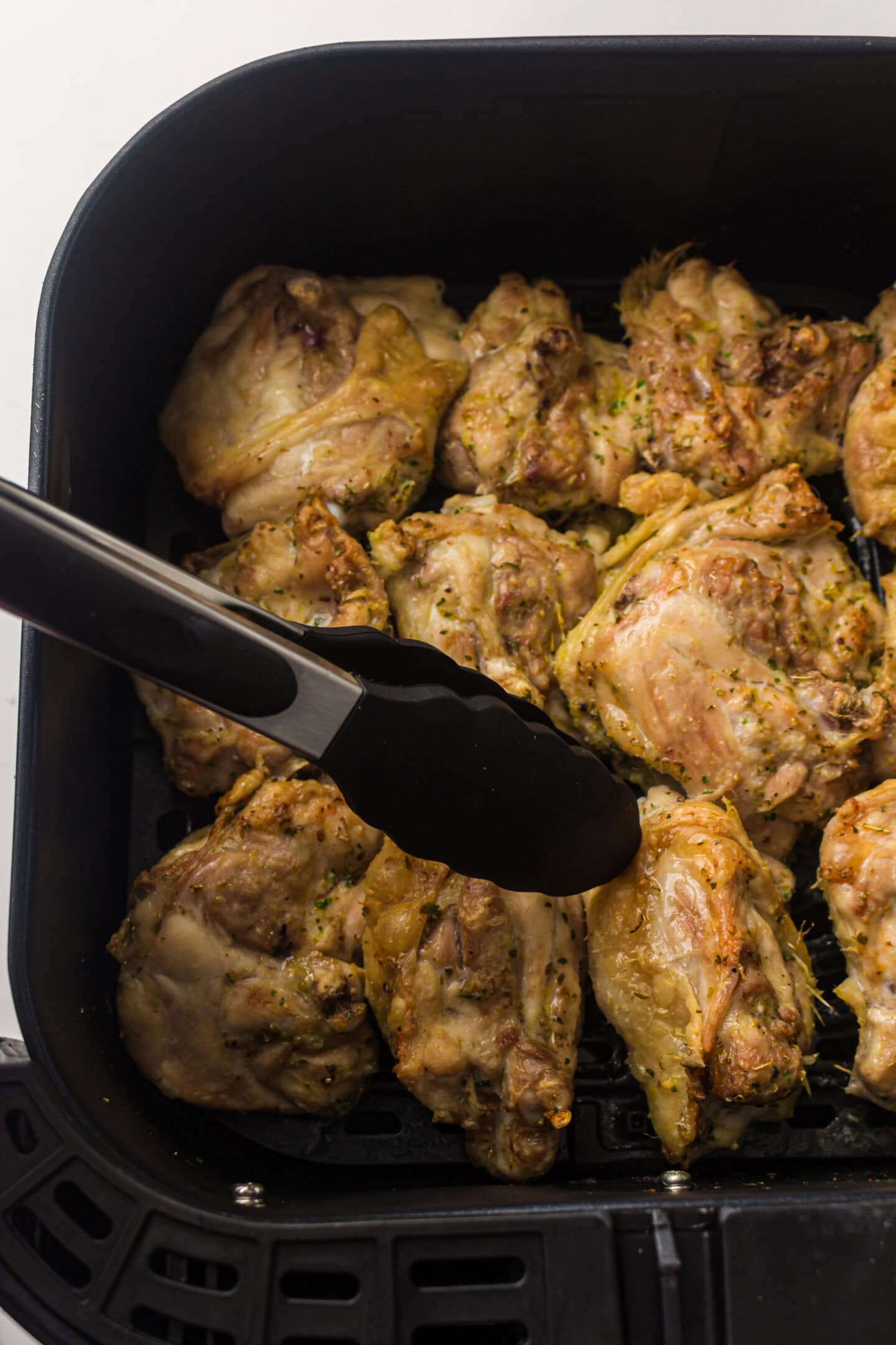 Chicken thighs cooked in an air fryer basket, with metal tongs holding one piece, shot from above on a white background.