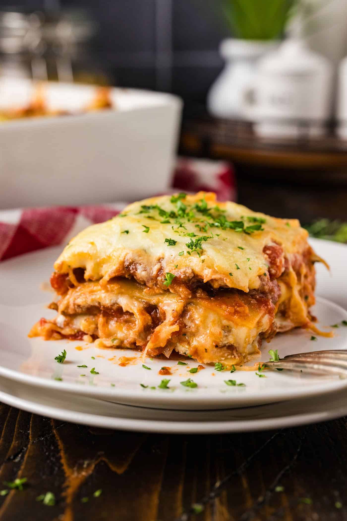 A slice of cheesy lasagna—one of the best Italian potluck ideas—topped with chopped parsley sits on a white plate with a fork; a baking dish is visible in the background.