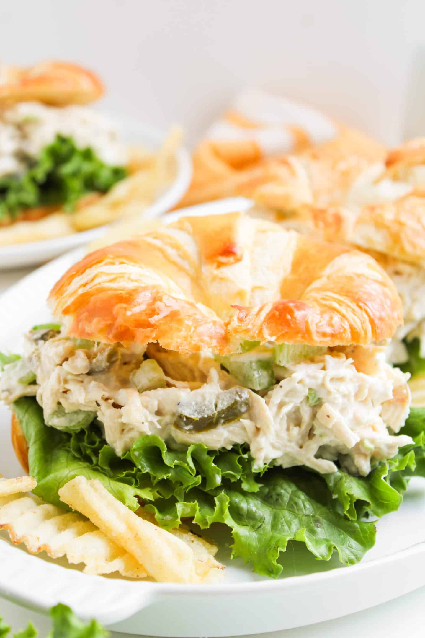 A croissant sandwich filled with Chicken Salad Chick Copycat chicken salad, lettuce, and pickles, served with crinkle-cut potato chips on a white plate.