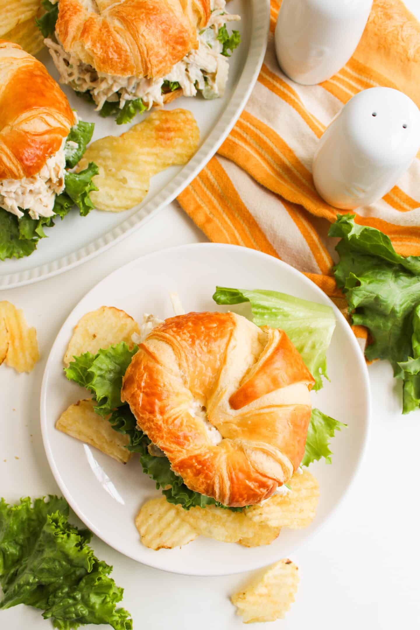 A croissant sandwich with lettuce and Jalapeño Holly Chicken Salad filling is served with potato chips on a white plate; a second sandwich is on a platter nearby with a salt and pepper set and an orange striped napkin for that Chicken Salad Chick copycat vibe.