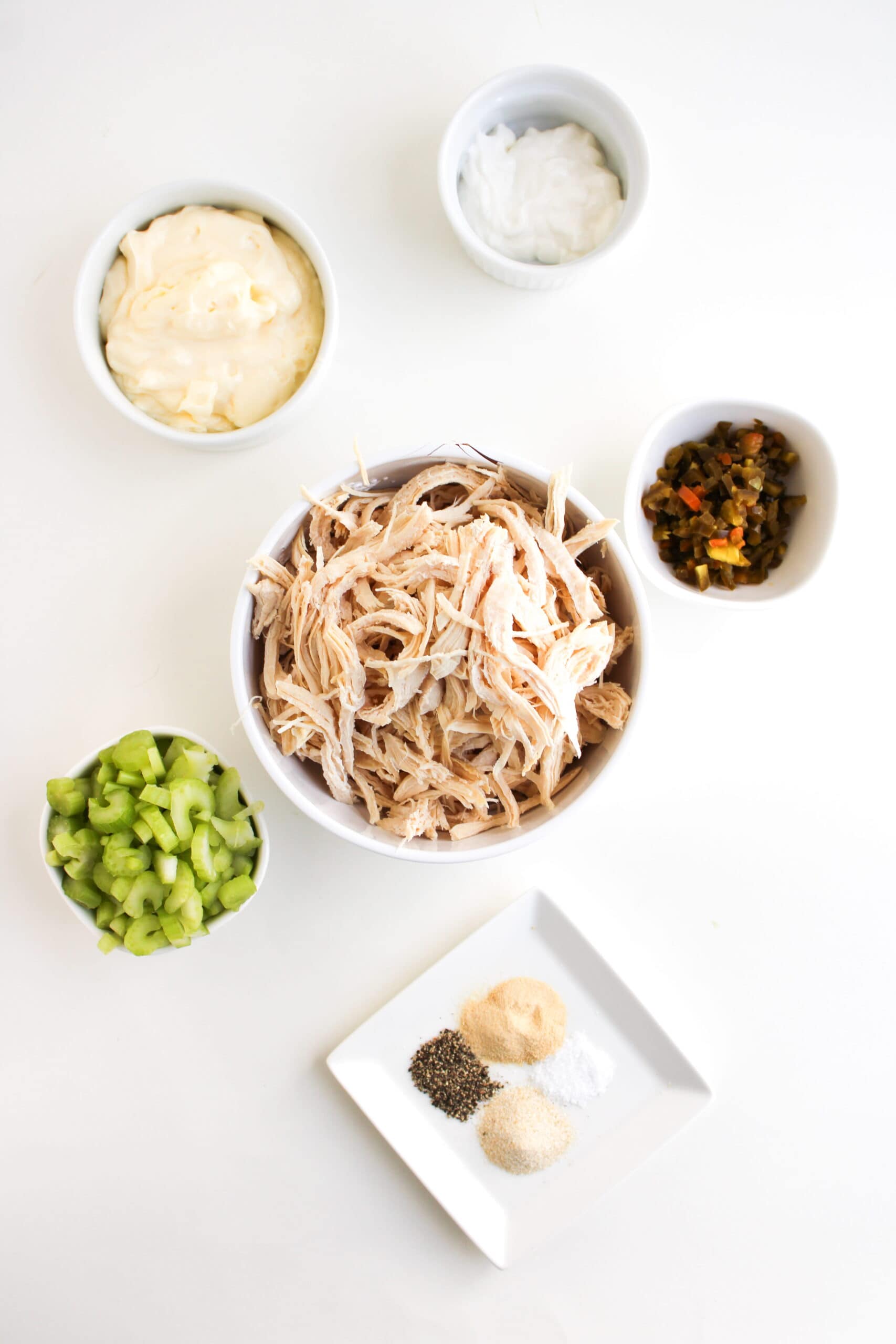 A bowl of shredded chicken is surrounded by small bowls of mayonnaise, yogurt, chopped celery, chopped pickles, and a plate of assorted spices, ready to create a Chicken Salad Chick Copycat or classic Jalapeño Holly chicken salad on a white surface.