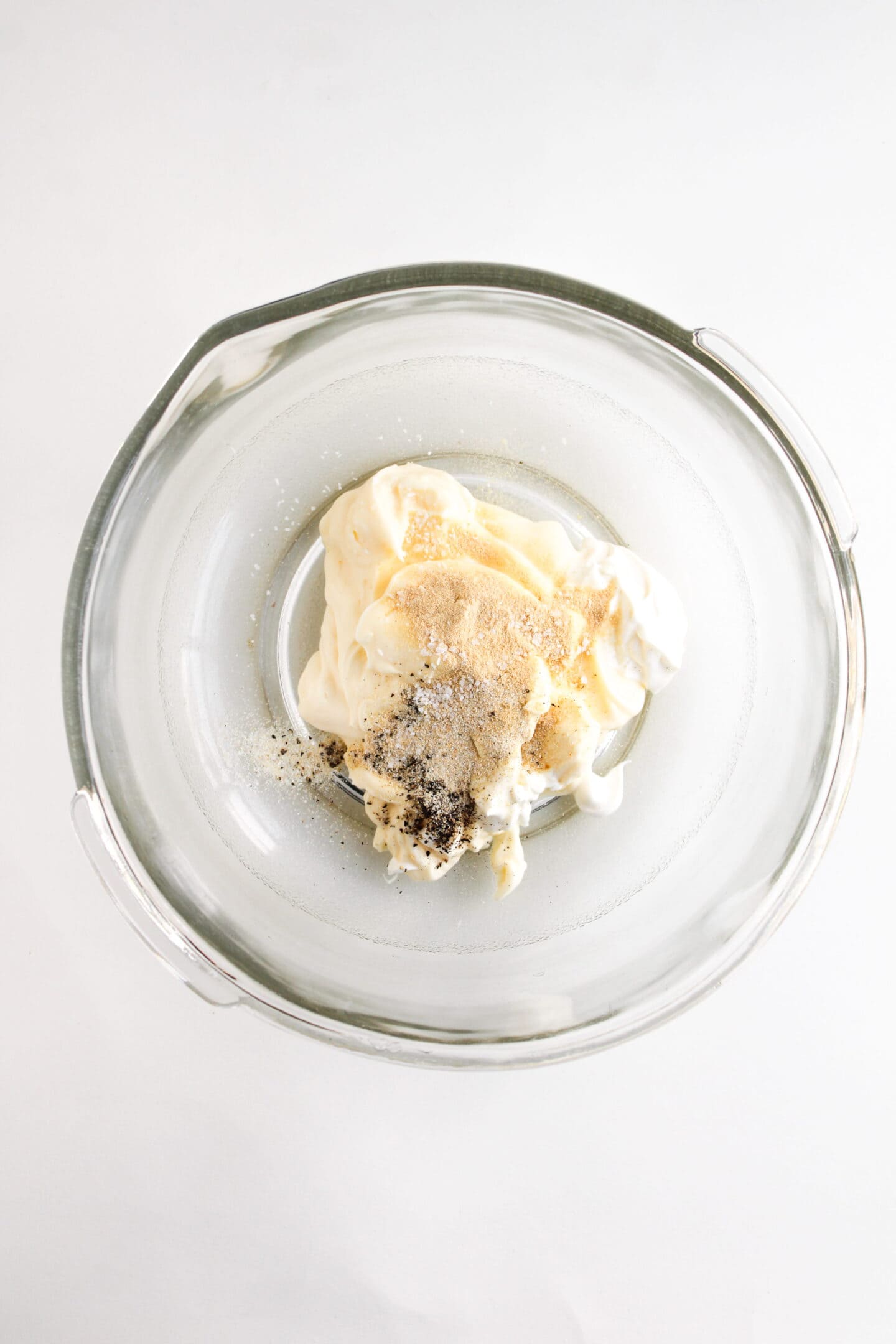 A glass bowl containing mayonnaise, sour cream, garlic powder, onion powder, and black pepper on a white surface—perfect for making a creamy Chicken Salad Chick Copycat.