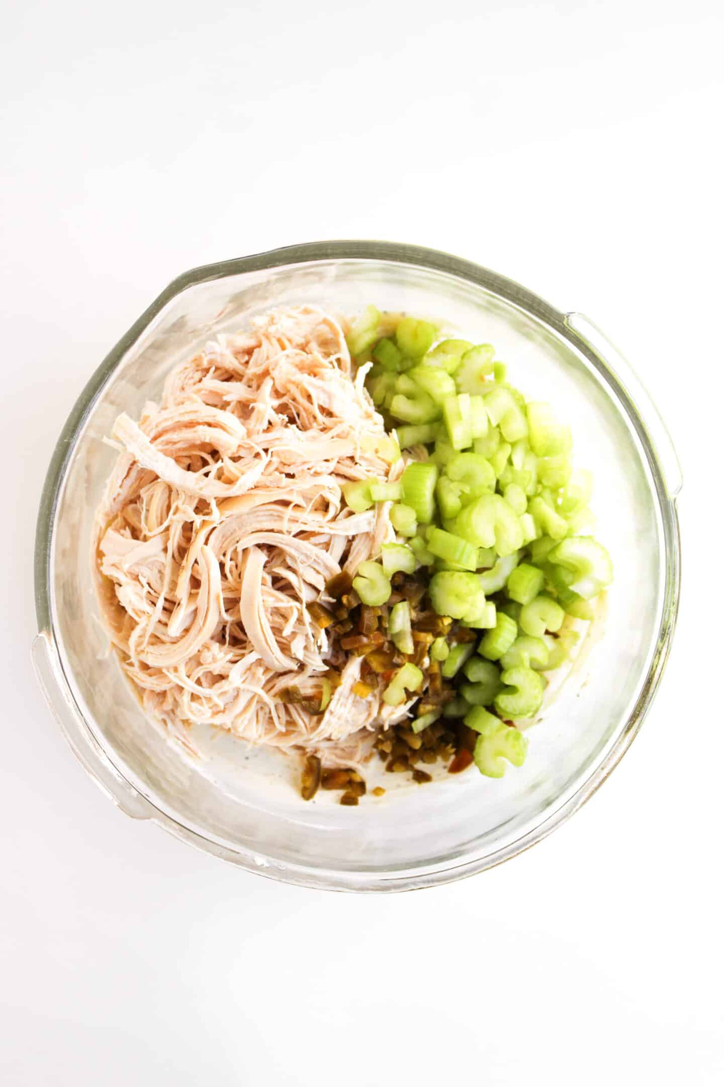 A glass mixing bowl containing shredded chicken, chopped celery, and chopped pickles—perfect for making a Jalapeño Holly Chicken Salad or your favorite Chicken Salad Chick Copycat recipe—sits on a white background.