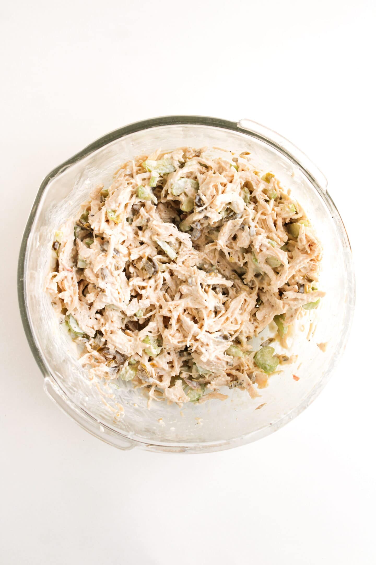 A clear glass bowl filled with shredded chicken salad, reminiscent of Chicken Salad Chick’s Jalapeño Holly Chicken Salad, mixed with mayonnaise, chopped celery, and other ingredients on a white surface.