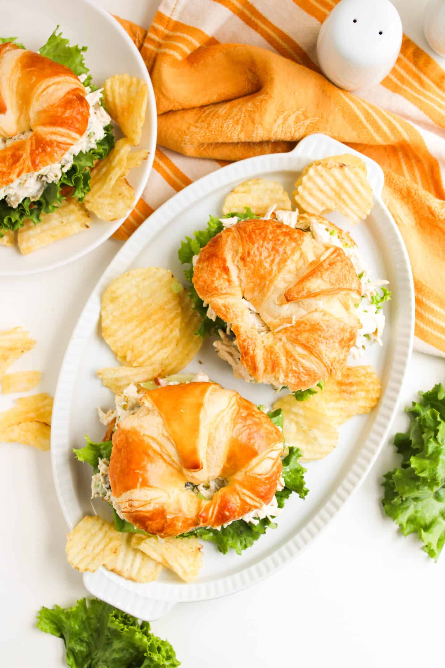Oval platter with two croissant sandwiches filled with Jalapeño Holly Chicken Salad and lettuce, served alongside crispy potato chips. An orange-striped towel and extra lettuce are in the background.