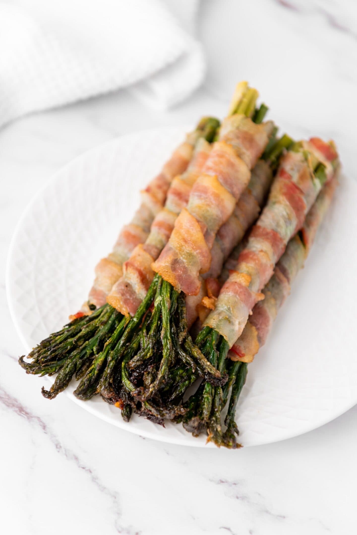 Three bundles of asparagus wrapped in bacon are arranged on a white plate on a marble surface, making them a perfect addition to your holiday side dishes or Easter recipes.