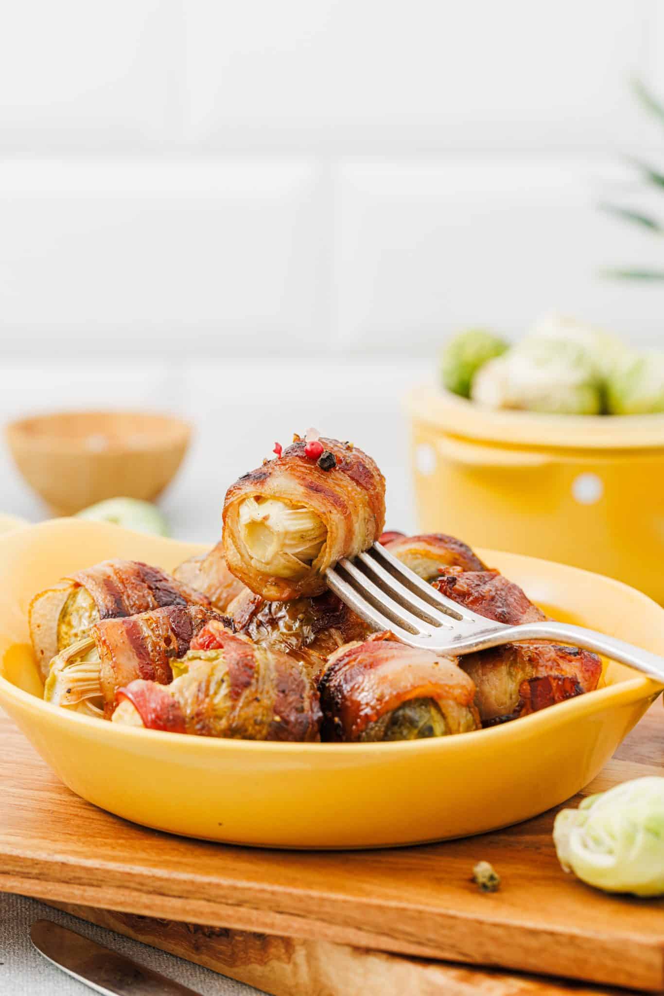 A yellow bowl filled with bacon-wrapped Brussels sprouts, with a fork holding one piece, set on a wooden board in a kitchen—perfect for adding to your Easter Side Dishes spread.