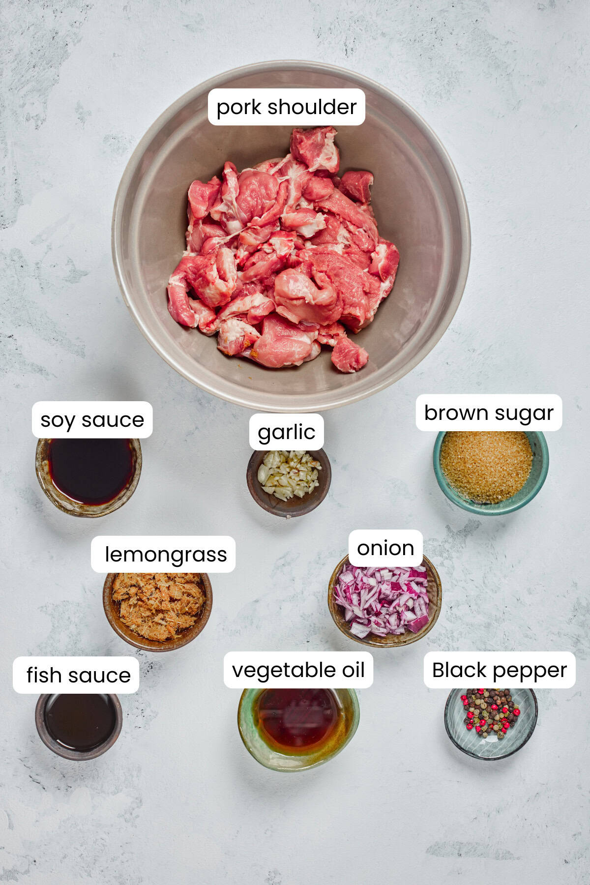 A bowl of sliced Vietnamese Grilled Pork shoulder is surrounded by small bowls of soy sauce, brown sugar, garlic, lemongrass, and more—perfect toppings for a classic Rice Noodle Bowl or Vietnamese Noodles.