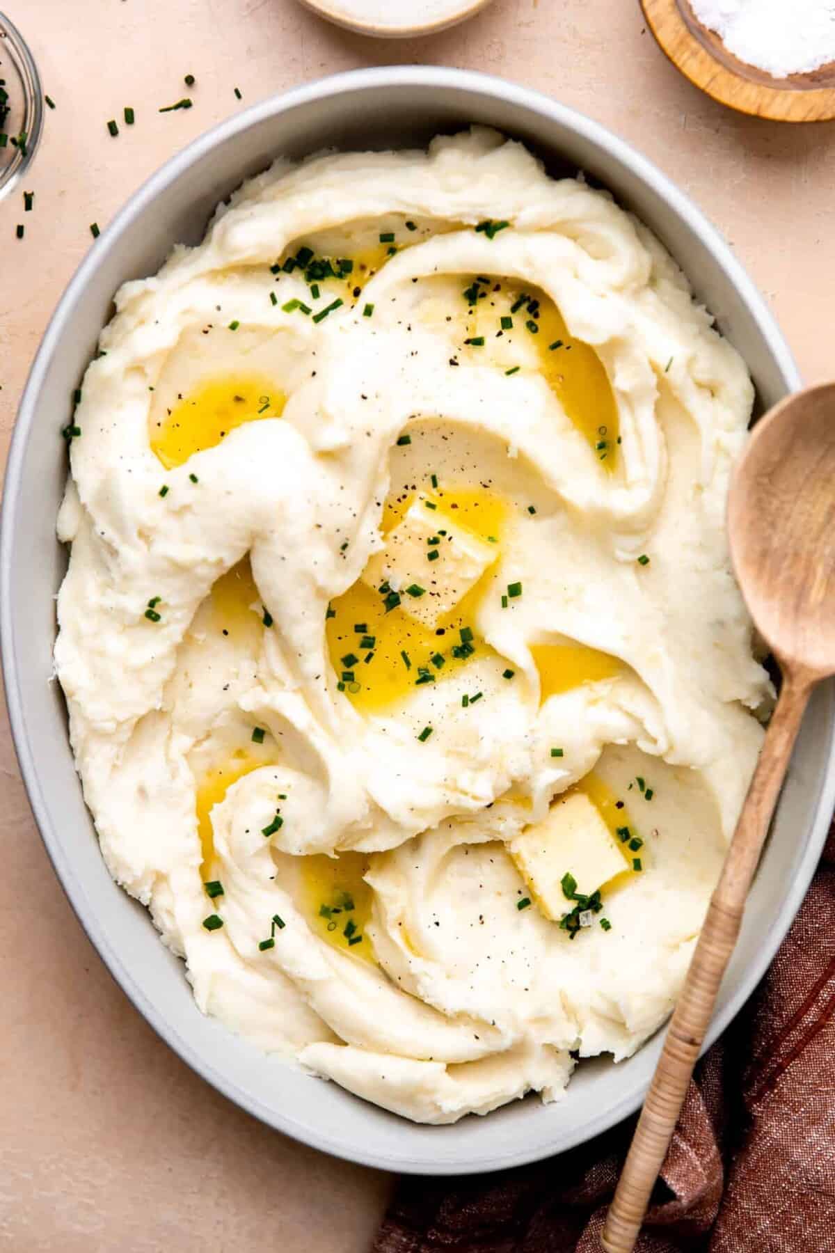 A bowl of mashed potatoes topped with melted butter and chopped chives, with a wooden spoon resting on the side—perfect for serving as one of your Easter side dishes.