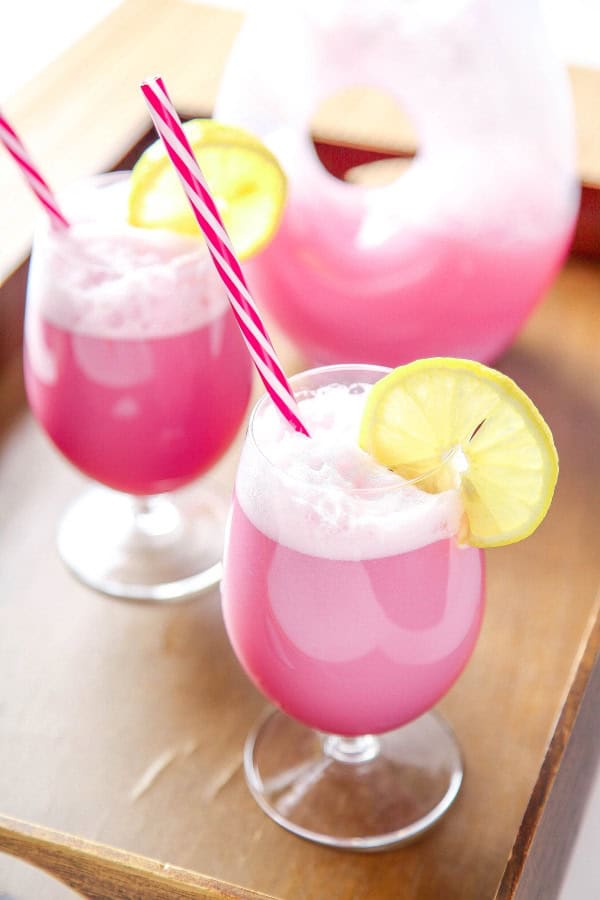 Two glasses of pink frothy drink with red-striped straws and lemon slices on the rim, placed on a wooden tray—perfect for your Easter Party Food spread. A matching pitcher sits in the background, ready to serve more festive refreshments.