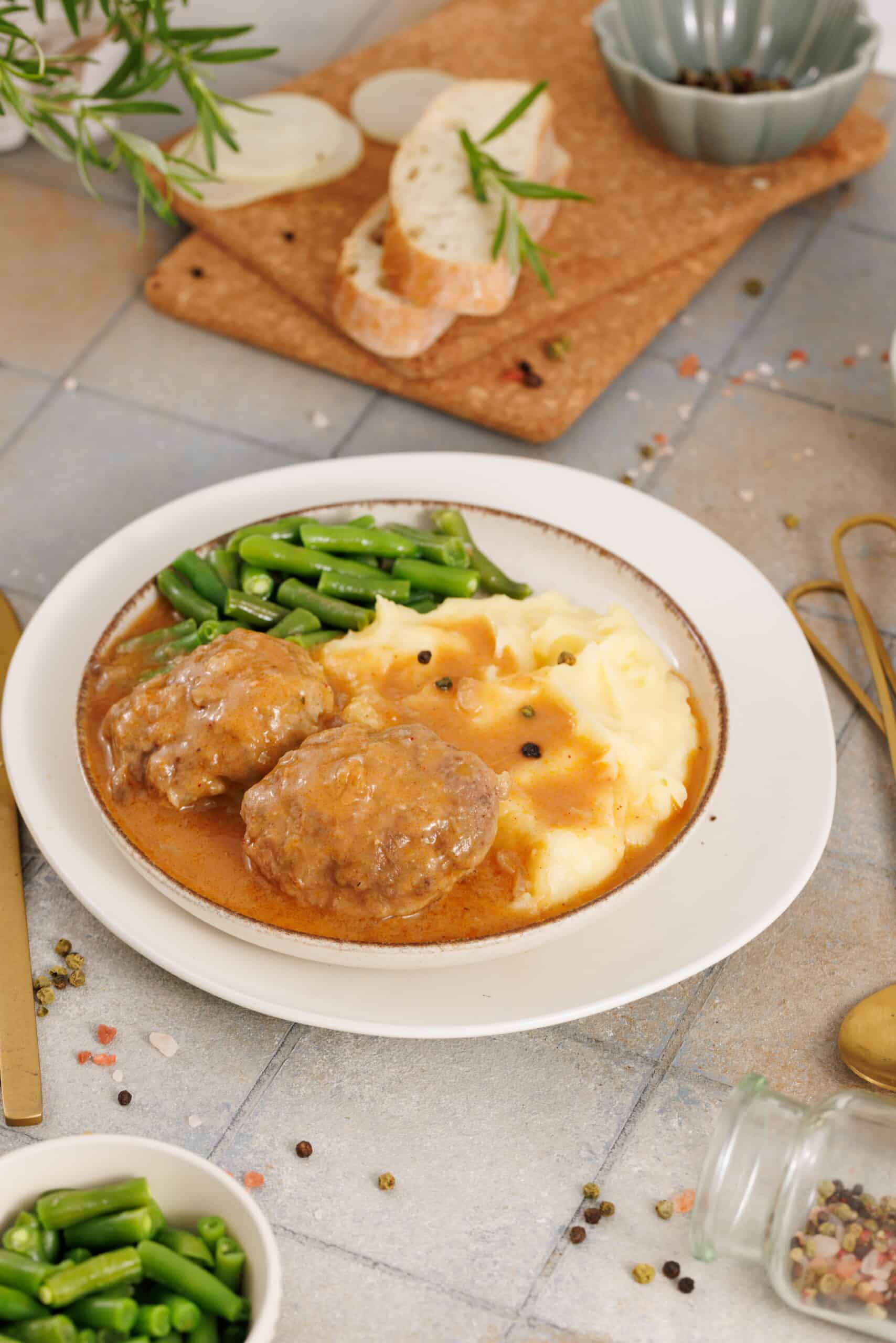 A plate of Salisbury Steak with Rich Brown Gravy, served with mashed potatoes, green beans, and sliced bread, with seasonings in the background.