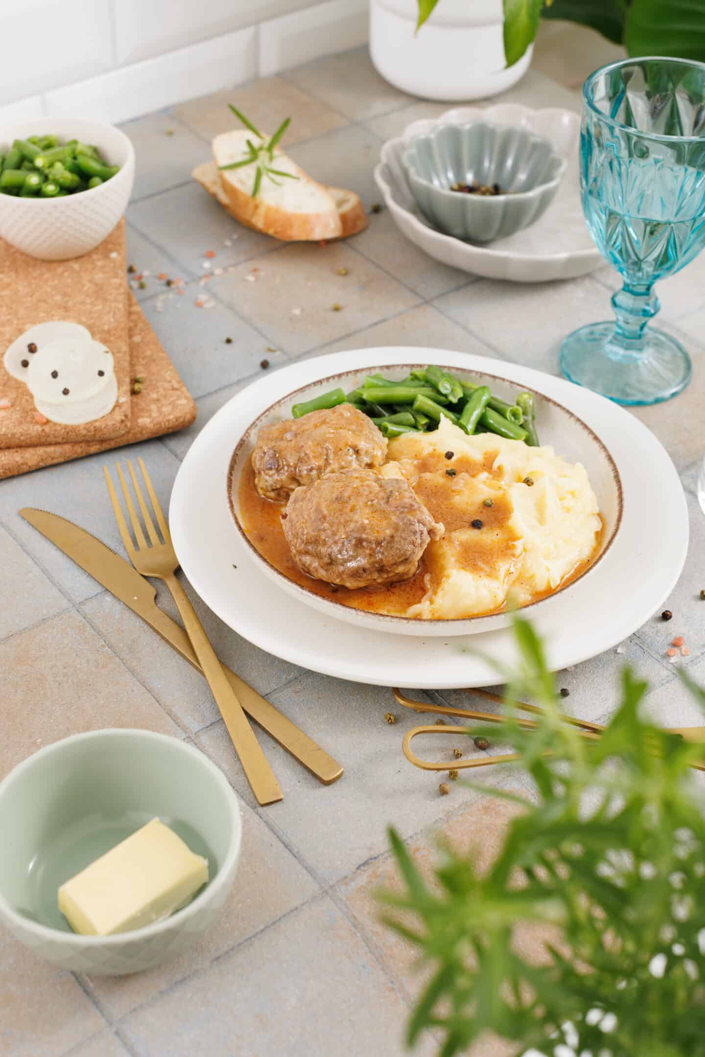 A plate with mashed potatoes, green beans, and Salisbury Steak with Rich Brown Gravy. Table setting includes gold utensils, a glass, butter, bread, and seasonings.