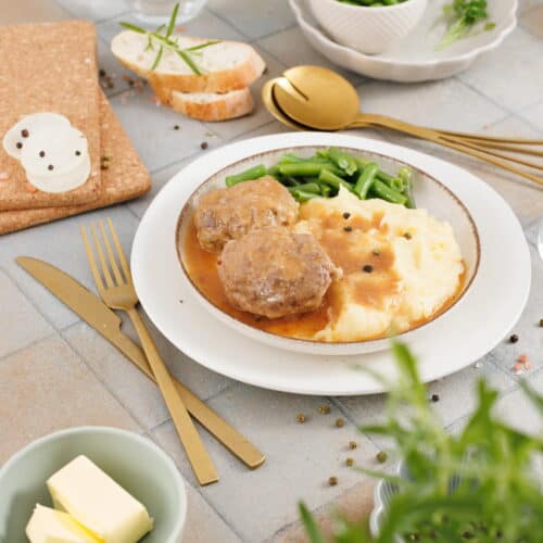 A plate with Salisbury Steak with Rich Brown Gravy, mashed potatoes, and green beans. Sliced bread, butter, extra green beans, and utensils are served on the side.