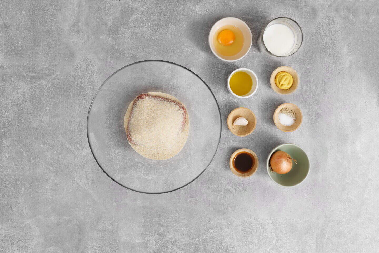 A glass bowl with raw ground meat topped with breadcrumbs, surrounded by small bowls containing ingredients for Salisbury Steak with Rich Brown Gravy, such as an egg, milk, oil, mustard, garlic, salt, pepper, and an onion on a gray surface.