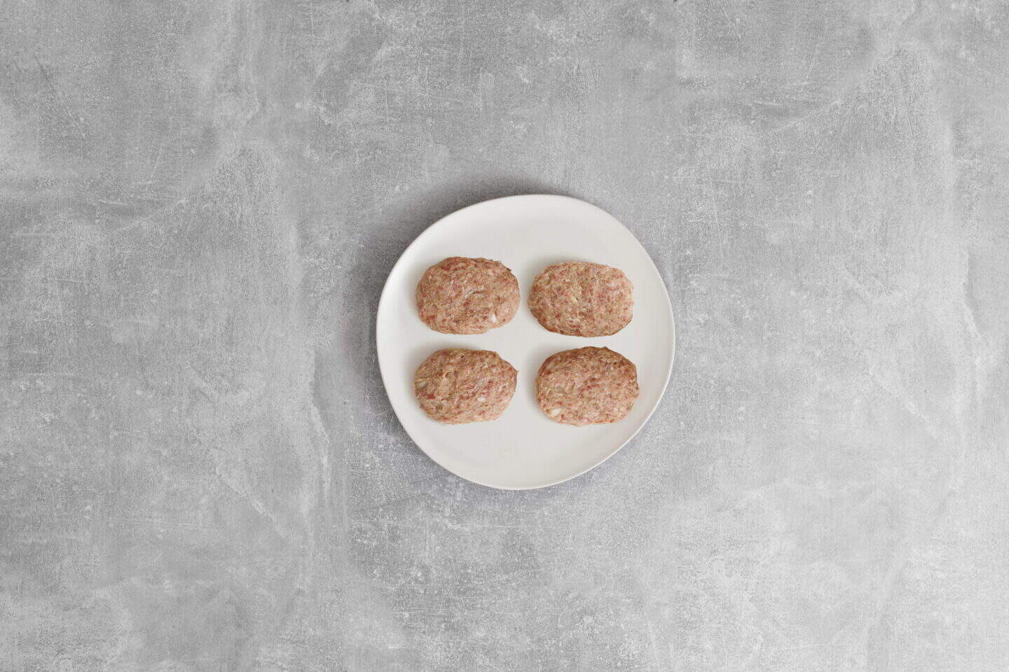 A white plate holds four raw, oval-shaped patties—perfect for making classic Salisbury Steak with Rich Brown Gravy—set on a gray textured surface.