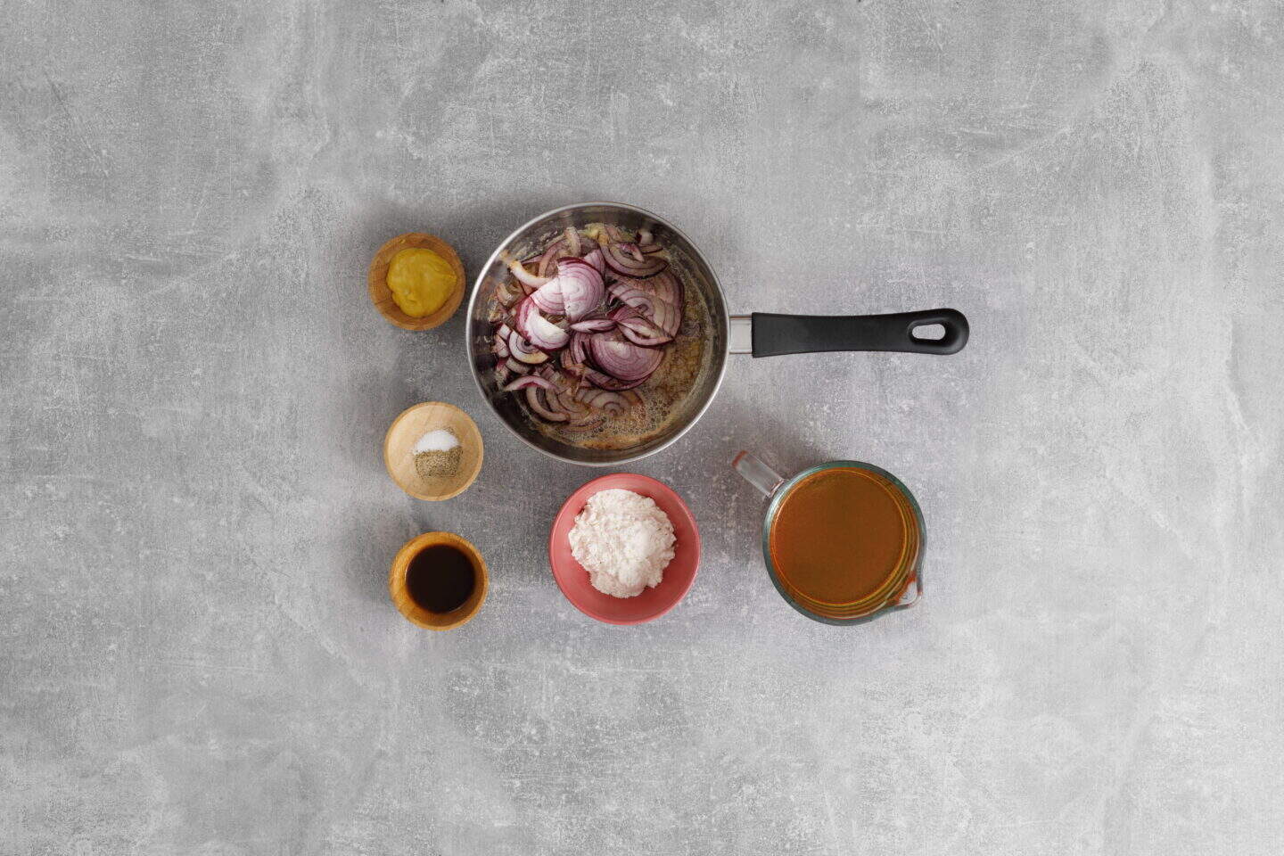 A saucepan with sliced red onions and five small bowls containing flour, broth, soy sauce, salt, and mustard—essential ingredients for preparing Salisbury Steak with Rich Brown Gravy—are arranged on a gray surface.